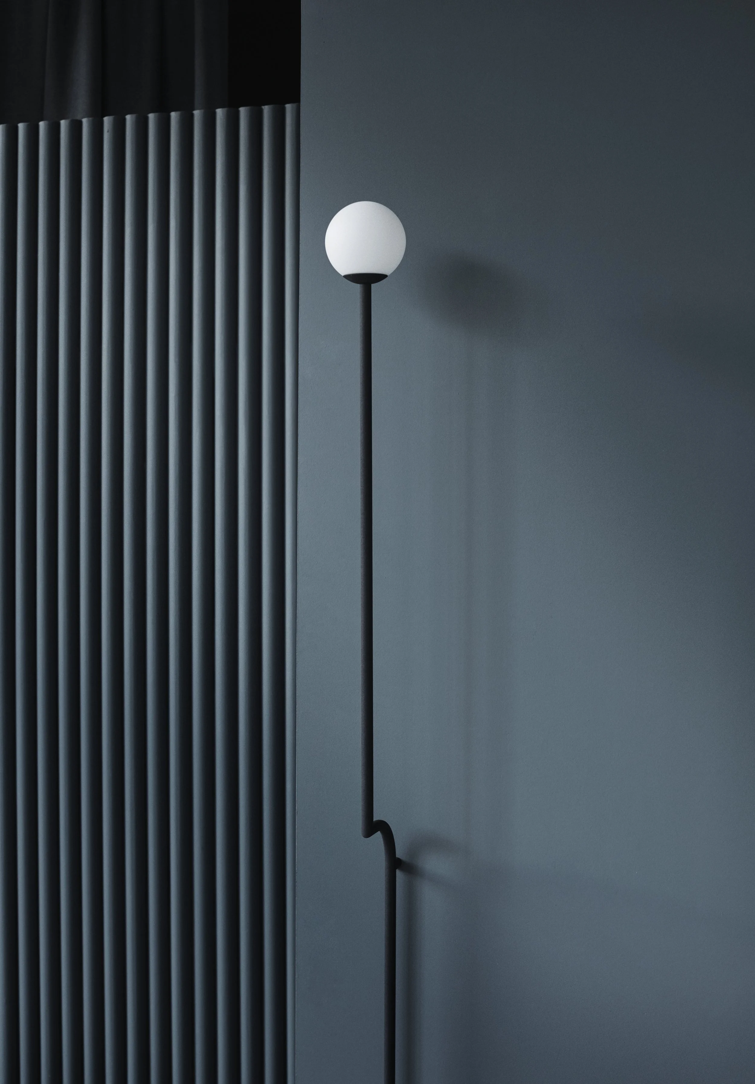 Modern black and white floor lamp with a spherical light on a black pole, positioned against a blue wall with textured vertical panels.