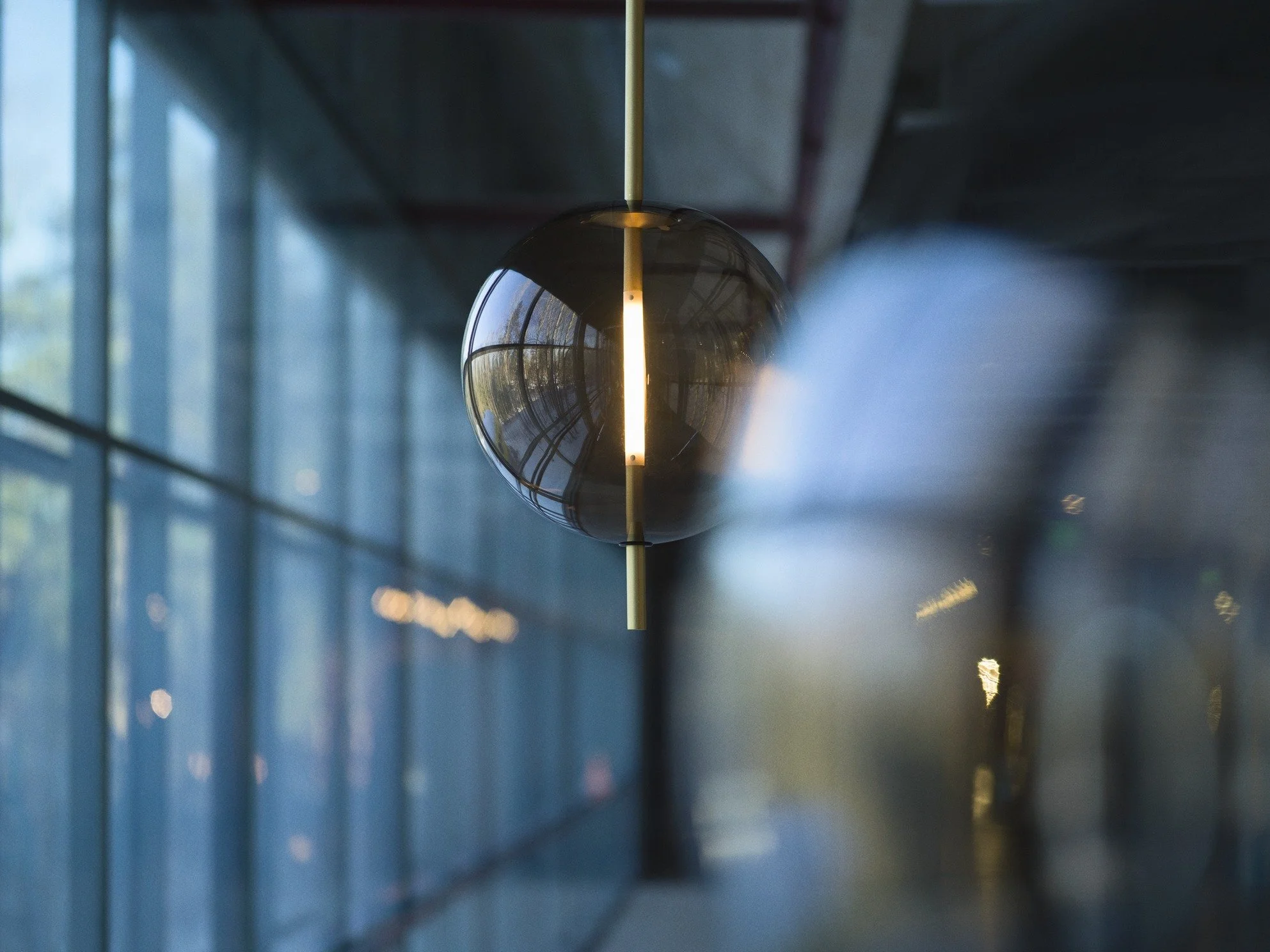 Close-up of a hanging black spherical pendant light in a modern building with blue-tinted glass walls and reflections.