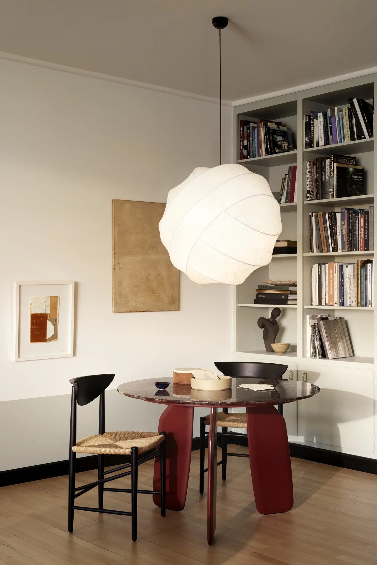 A modern dining room featuring a round table with a curved red base, black marble top, and two black chairs with woven seats. A large white paper pendant light hangs from the ceiling. The background includes a white wall with abstract art and a tall 