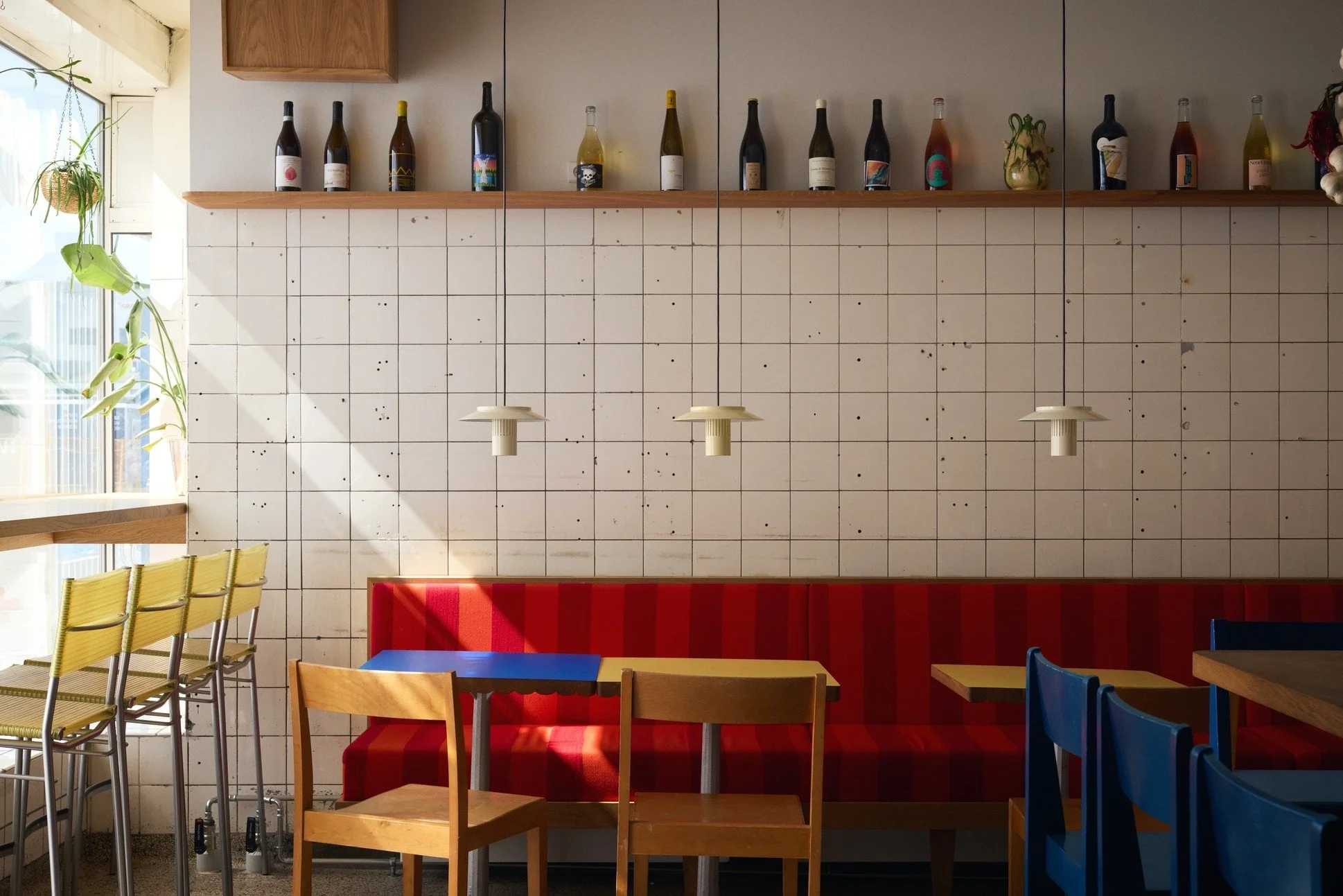 Interior of a restaurant with a tiled wall, red cushioned bench seating, and chairs in yellow, natural wood, and blue. Bottles are on a shelf above the wall, and hanging pendant lights are above the tables. A window with plants is on the left.
