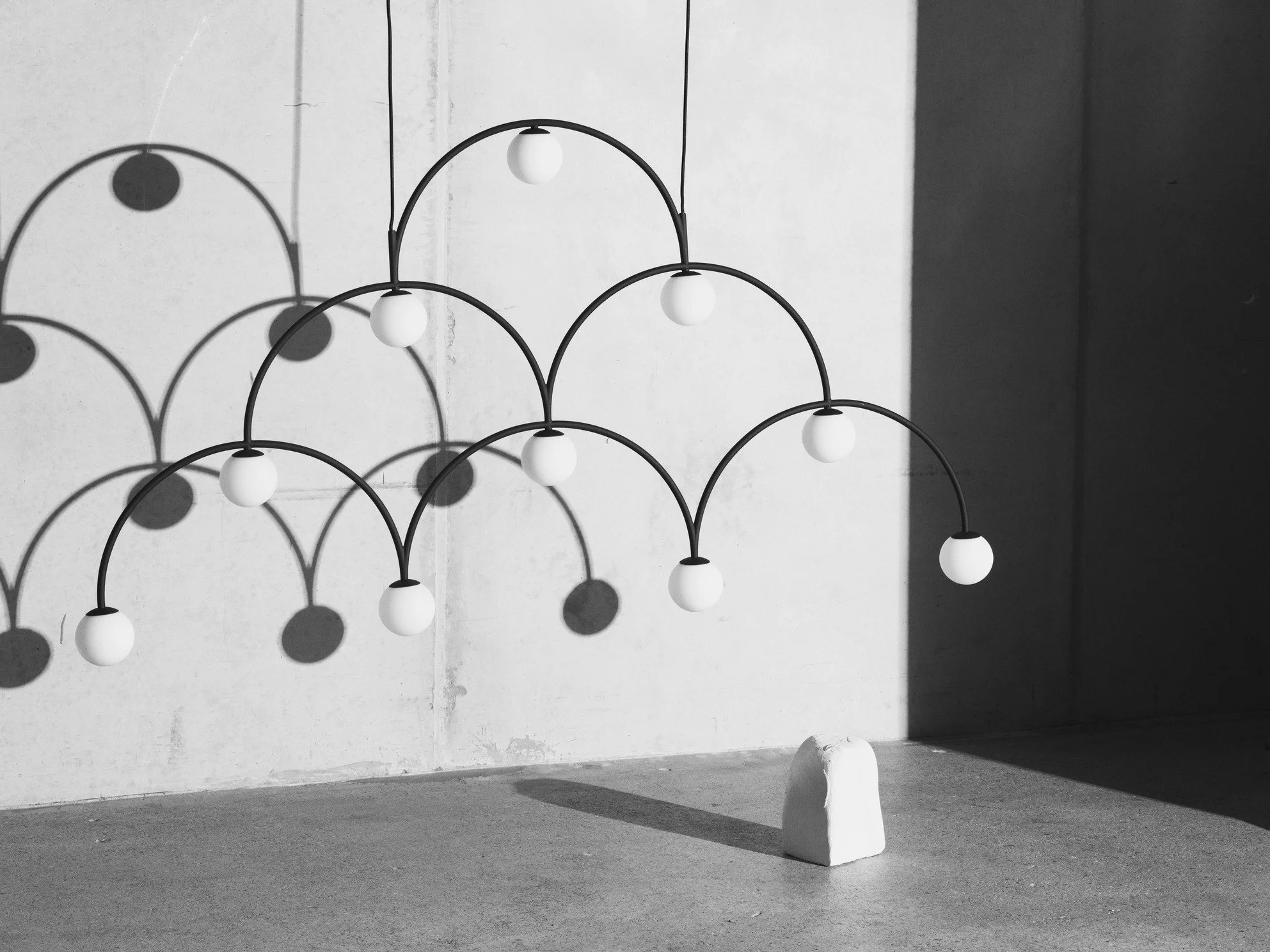 Modern chandelier with multiple curved arms holding round white light bulbs, casting shadows on the wall behind it in a minimalist space.