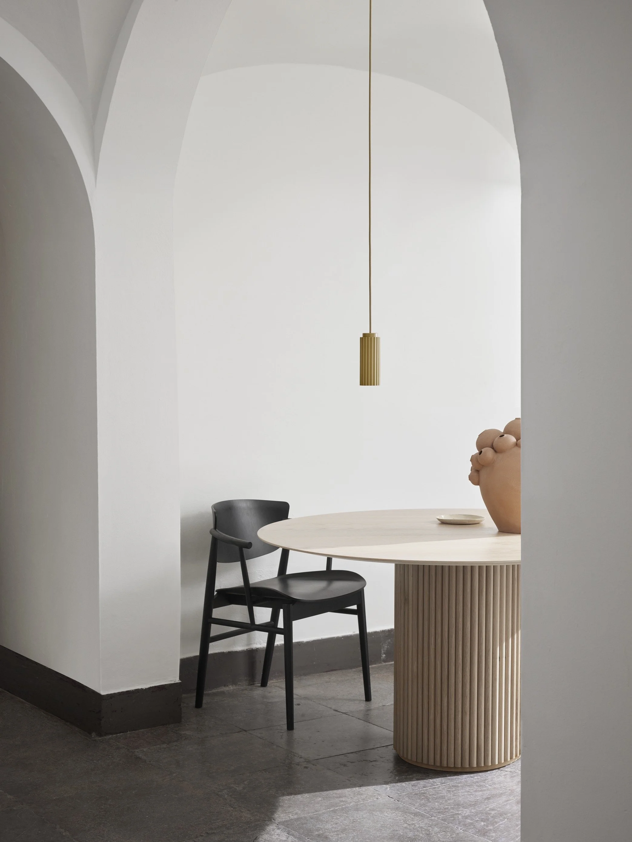 Minimalist interior with a round wooden table, a black chair, a hanging pendant light, and a large pottery vase with smaller round shapes on top of it.