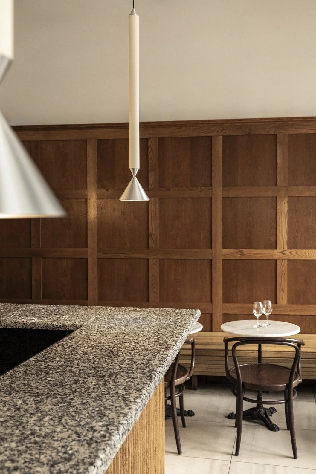 Interior of a restaurant with a granite countertop in the foreground and a small round table with two glasses on it in the background. The wall is wooden paneling.