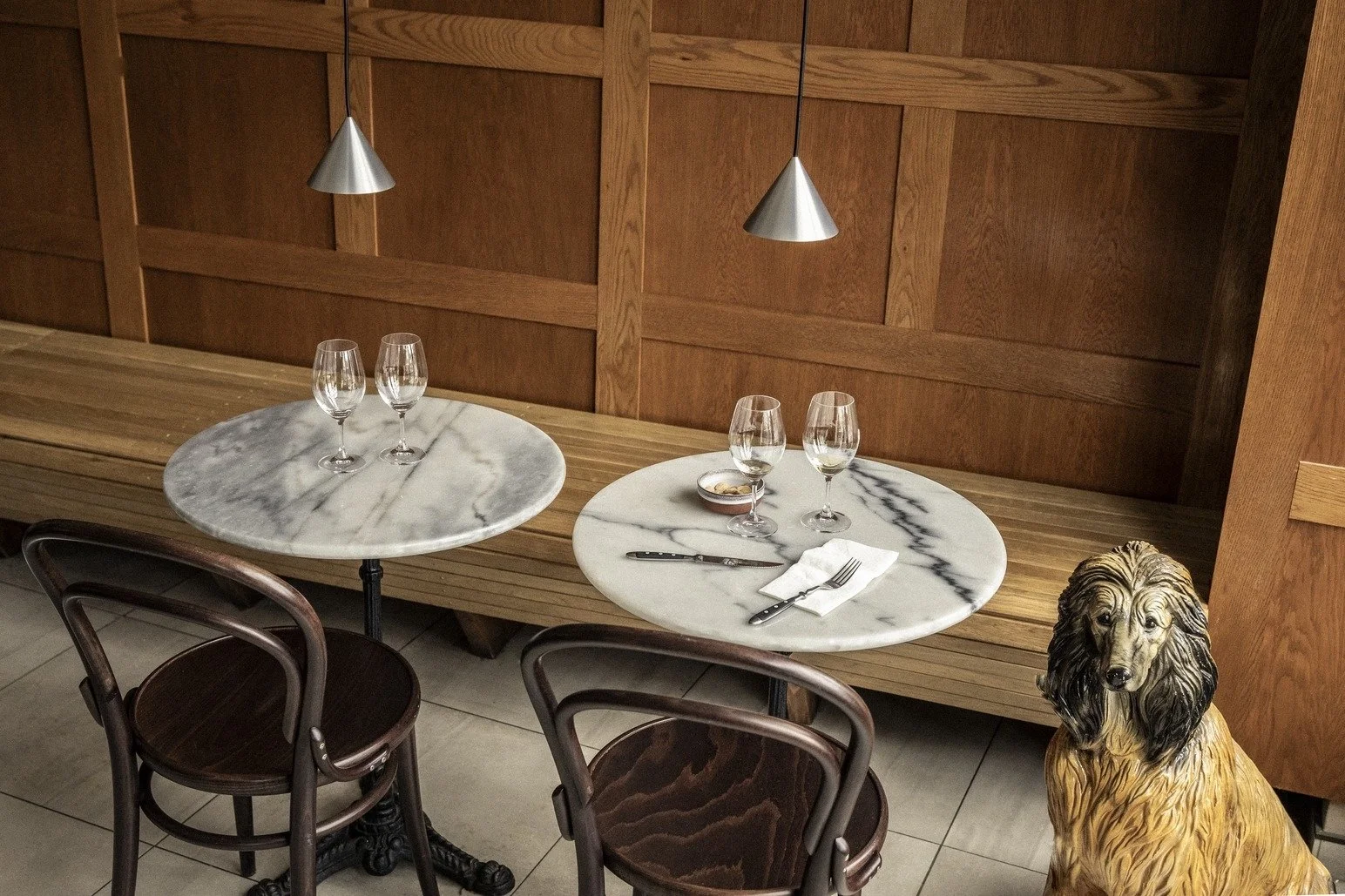 A cozy restaurant table set with four wine glasses, a small bowl, and utensils on marble tabletops, with a wooden bench and wall decor, and a ceramic dog statue in the foreground.