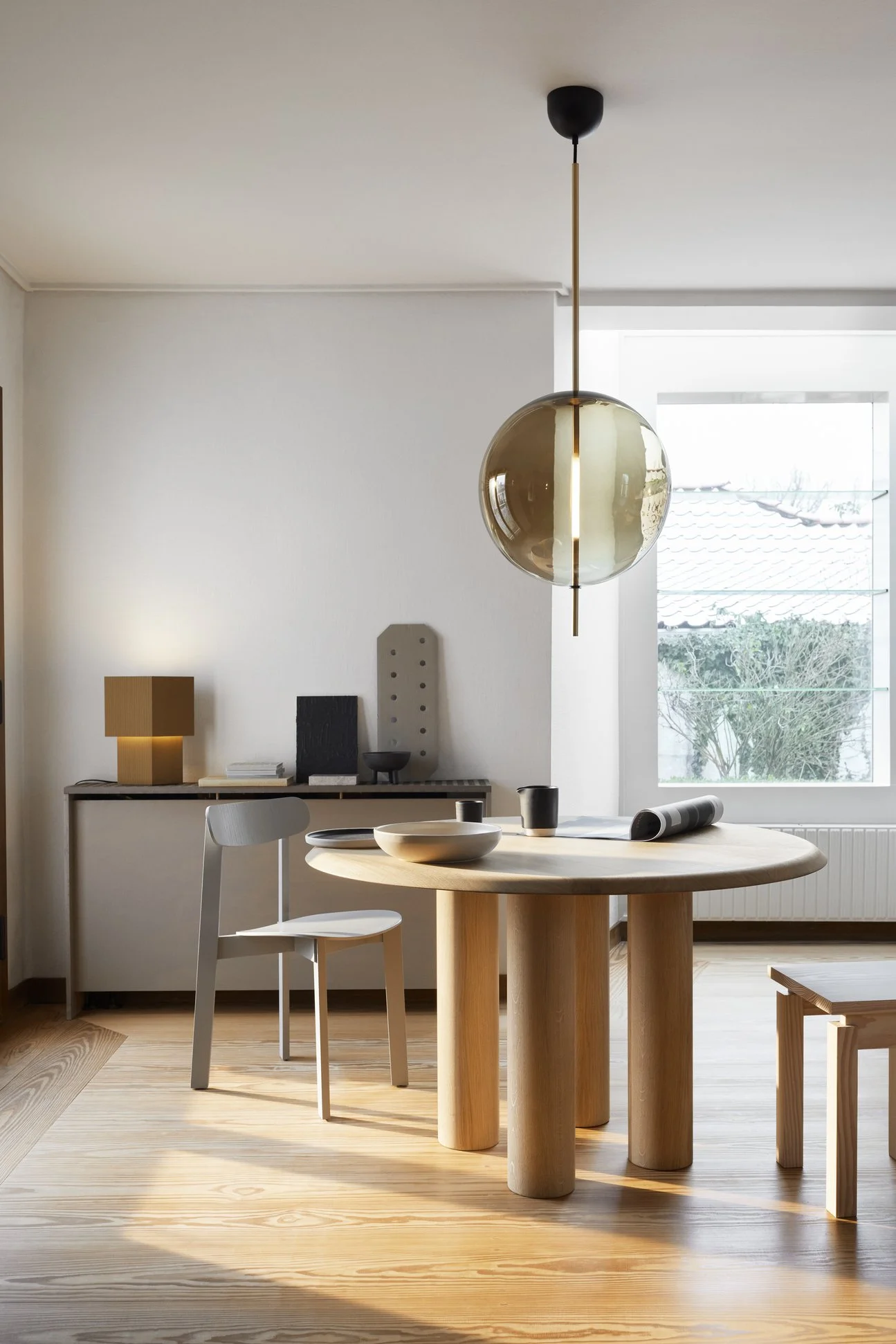 A minimalistic dining room with a round wooden table, modern white and black chairs, decorative objects on a sideboard, and a large window letting in natural light.