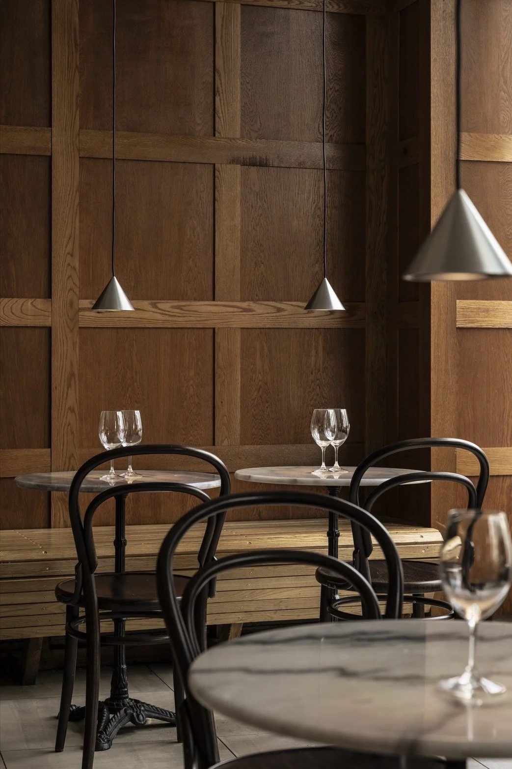 Empty restaurant with wooden wall paneling, black chairs, marble-topped tables, and hanging cone-shaped pendant lights.