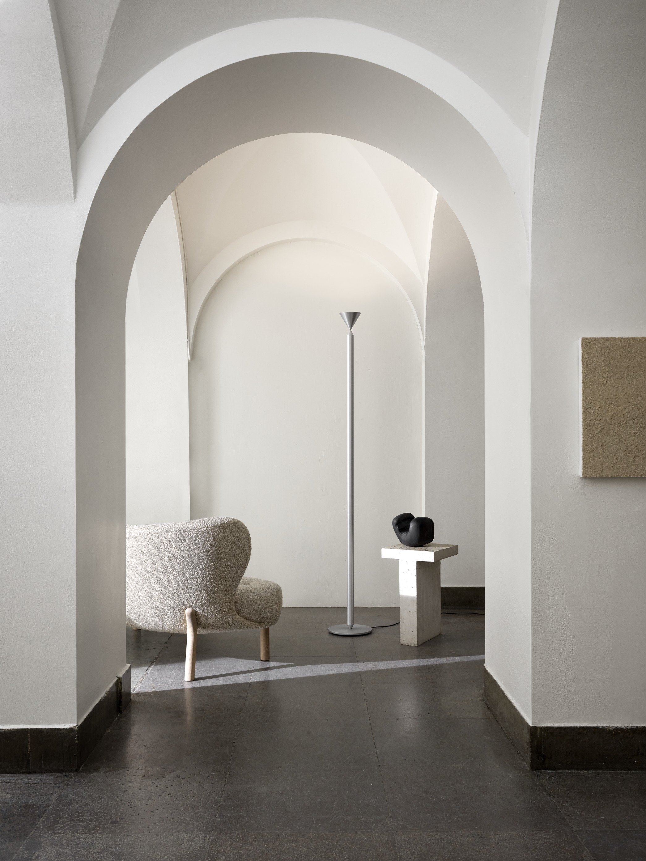 Minimalist room with white arched ceiling, a textured beige armchair, a tall floor lamp, and a small pedestal with a black abstract sculpture.