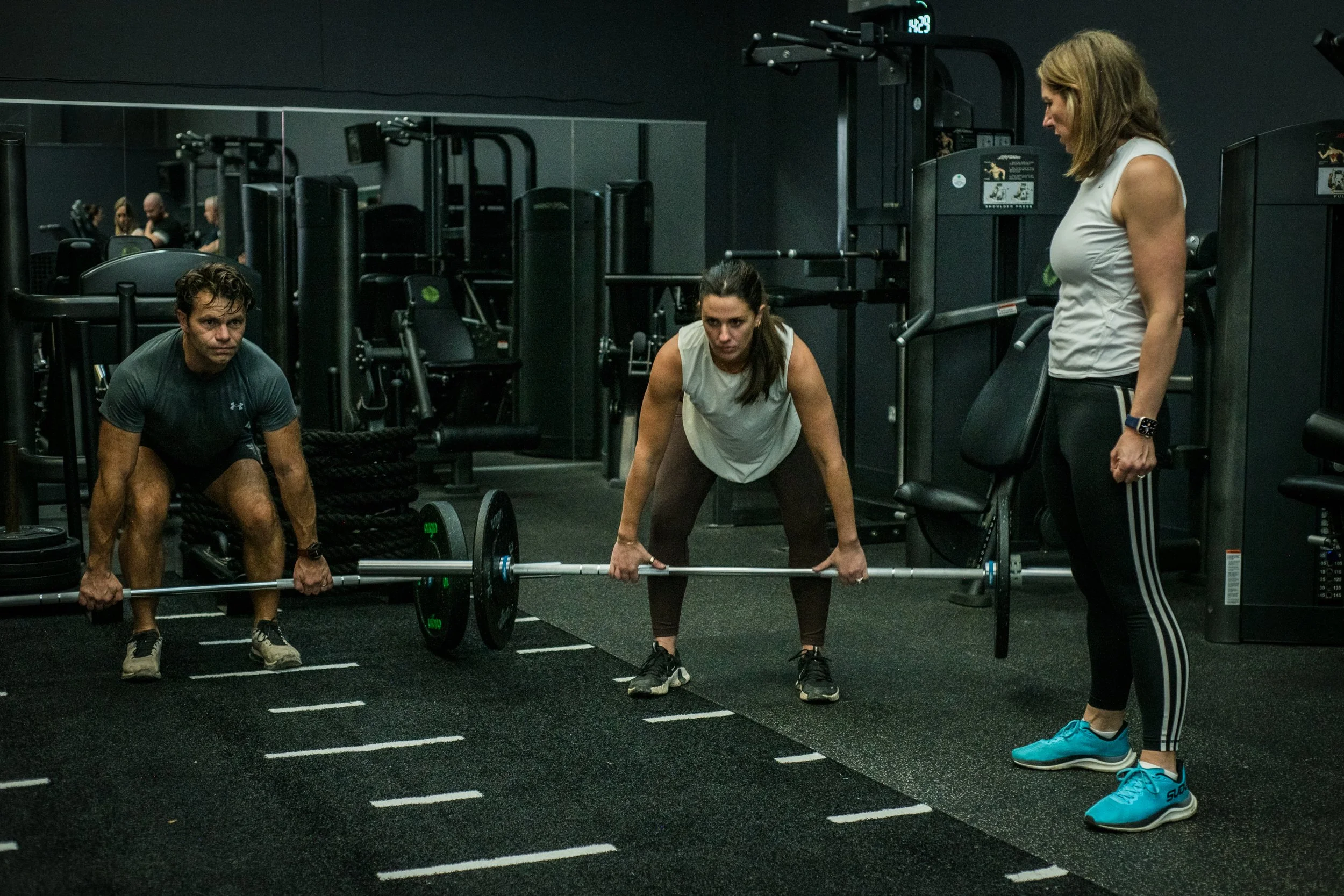 A woman is preparing to lift a barbell, supervised by a trainer, in a gym with two other individuals in workout attire and various gym equipment in the background.