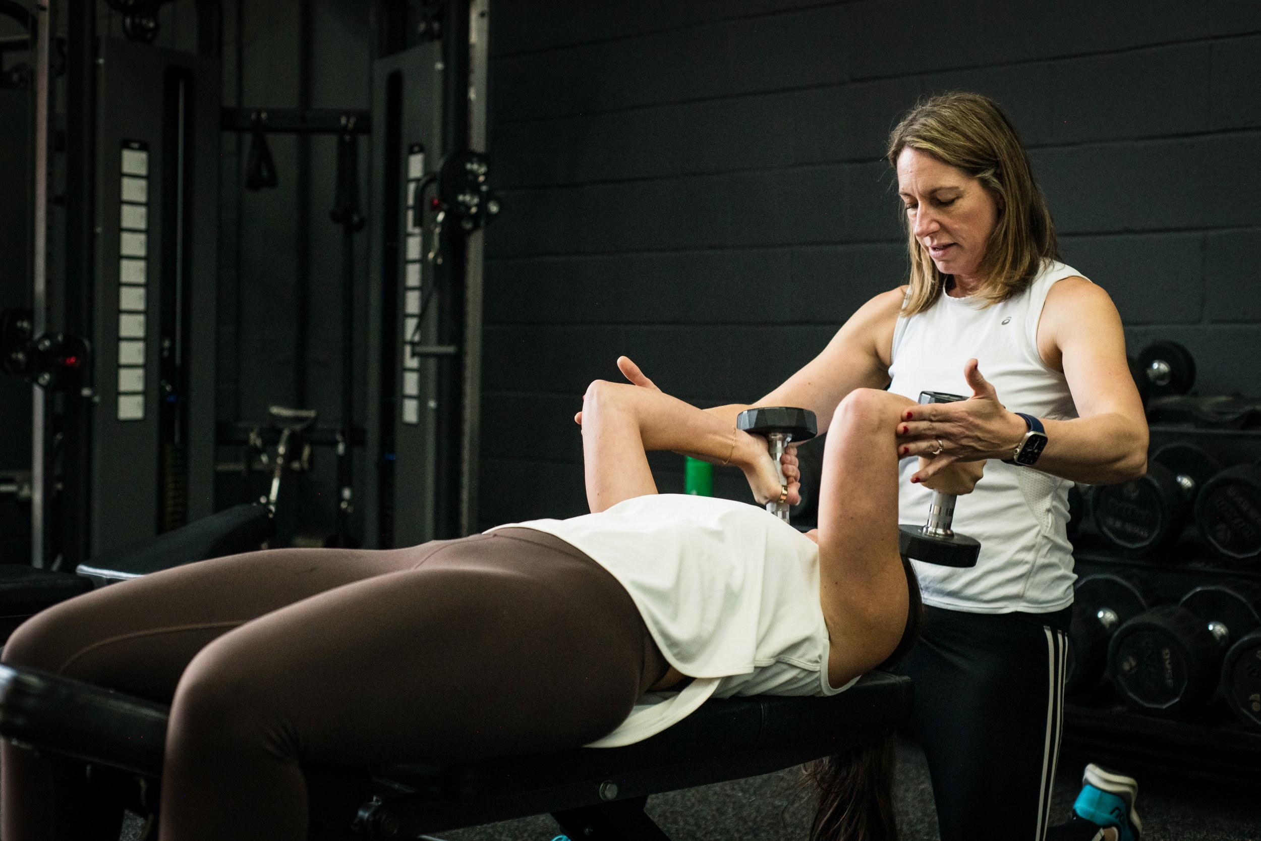 Personal trainer assisting woman with dumbbell exercise in gym.