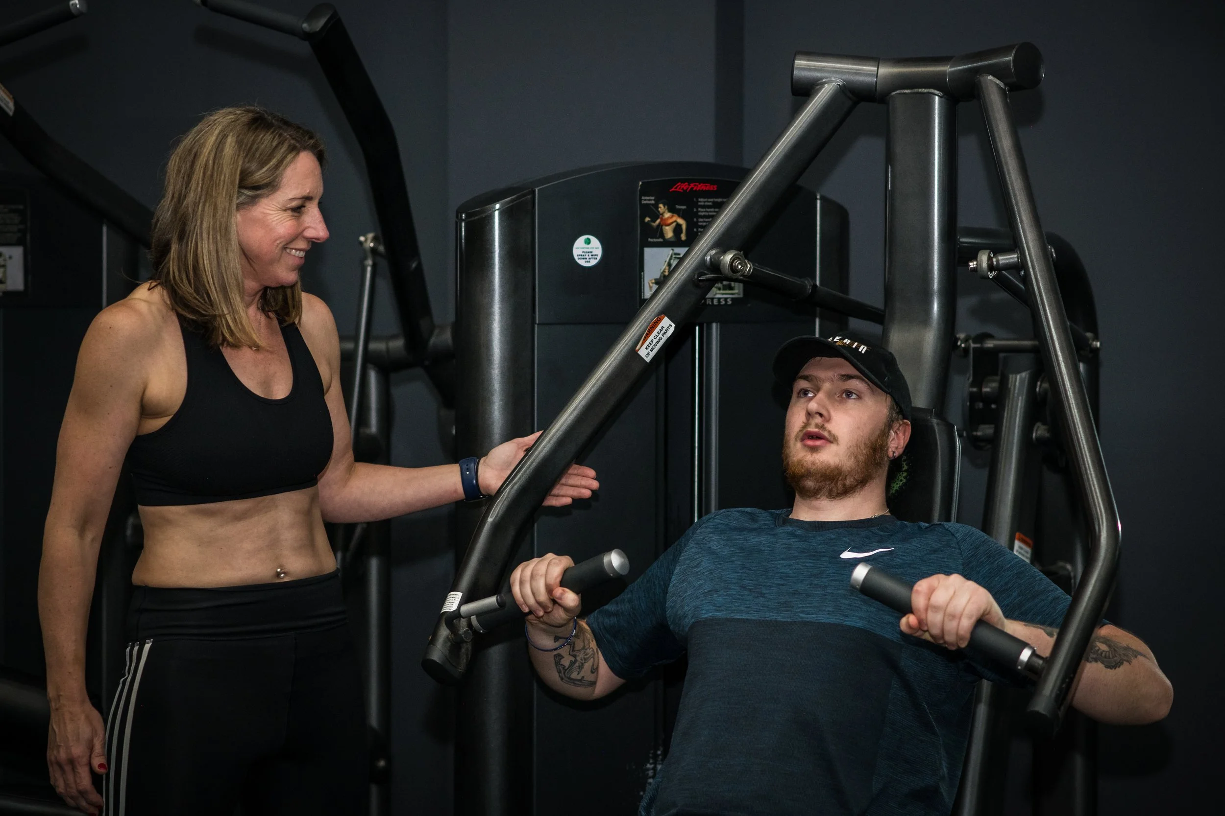 A woman in a black sports bra and black pants assisting a man in a dark blue athletic shirt with a workout at a gym. The man is using a chest press machine, and the woman is standing beside him, offering support and encouragement.