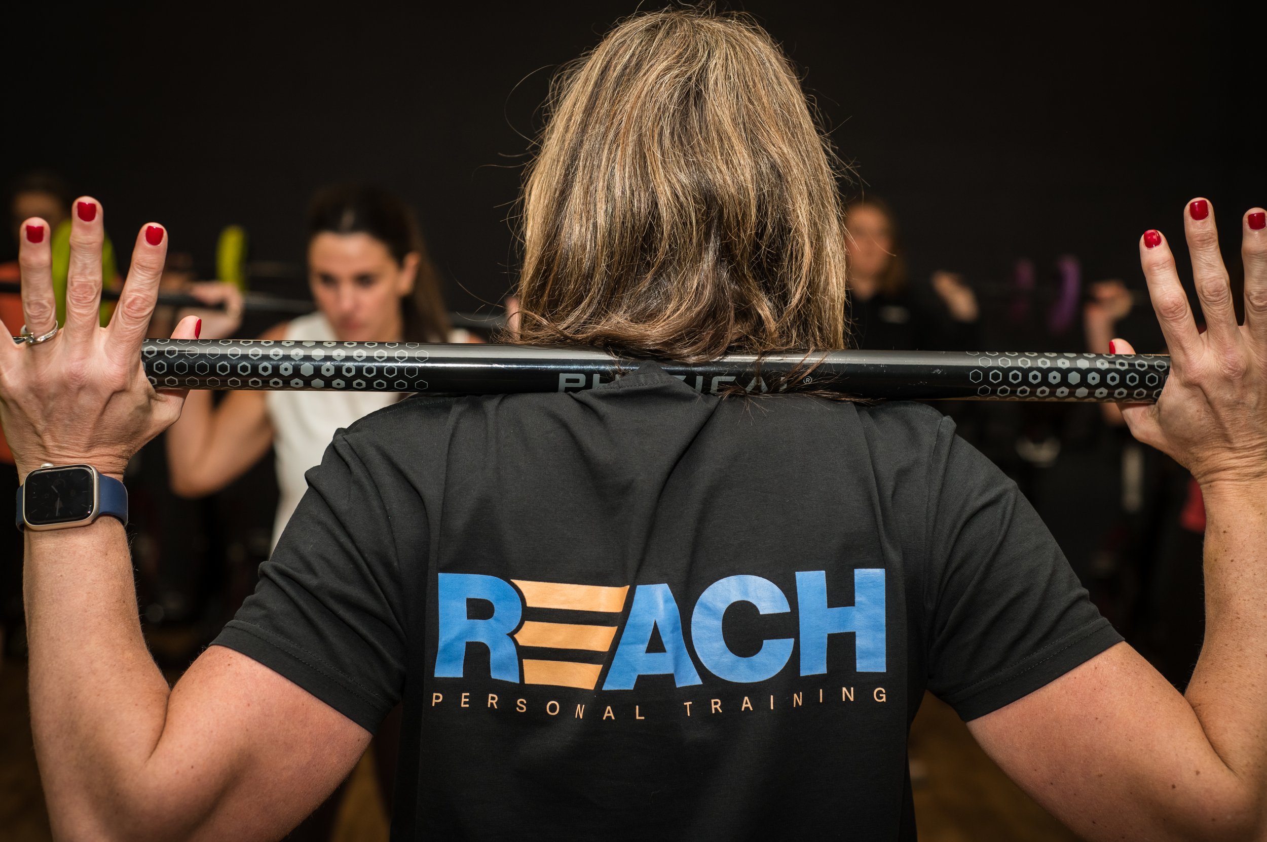 Rear view of a woman in a black T-shirt with the word 'REACH' and 'personal training' written on it, holding a barbell behind her neck during a fitness class in a gym with other participants in the background.