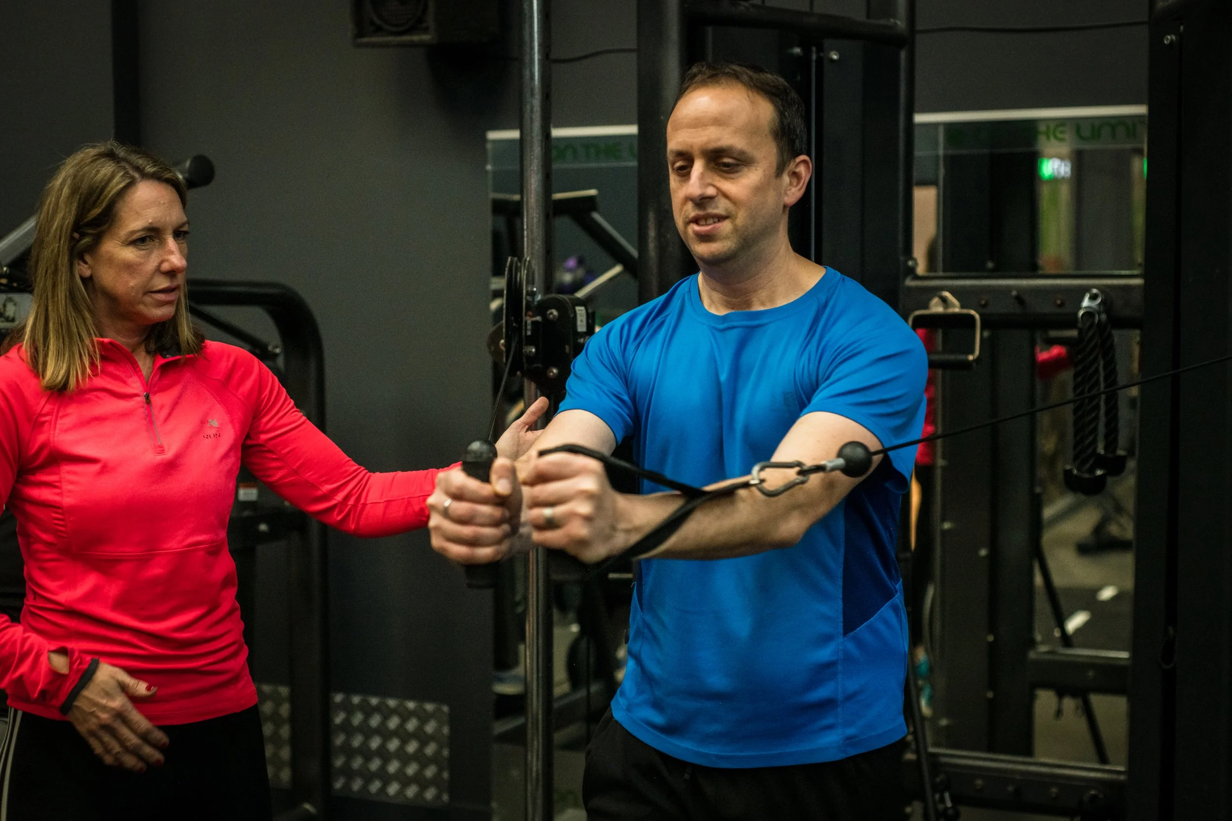 A man wearing a blue athletic shirt performs a resistance exercise with a trainer guiding him, in a gym setting.