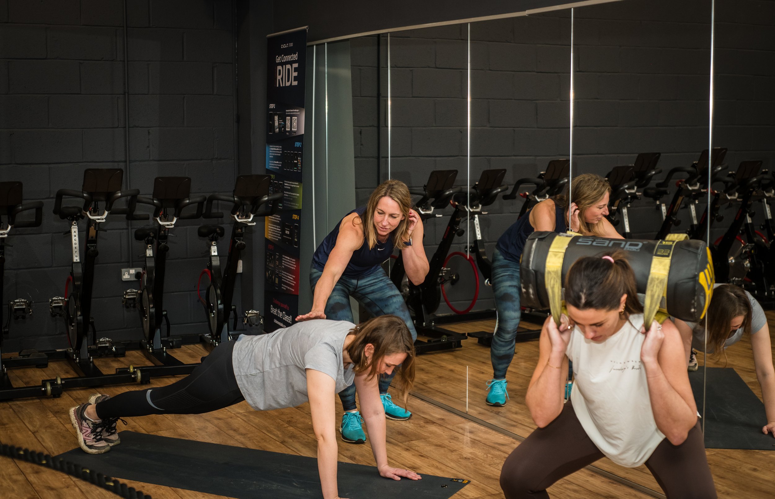 Women exercising in a gym, including a woman doing a plank, a woman doing a squat with a weight, and a woman doing a bent-over row, with a mirrored wall and bicycles in the background.