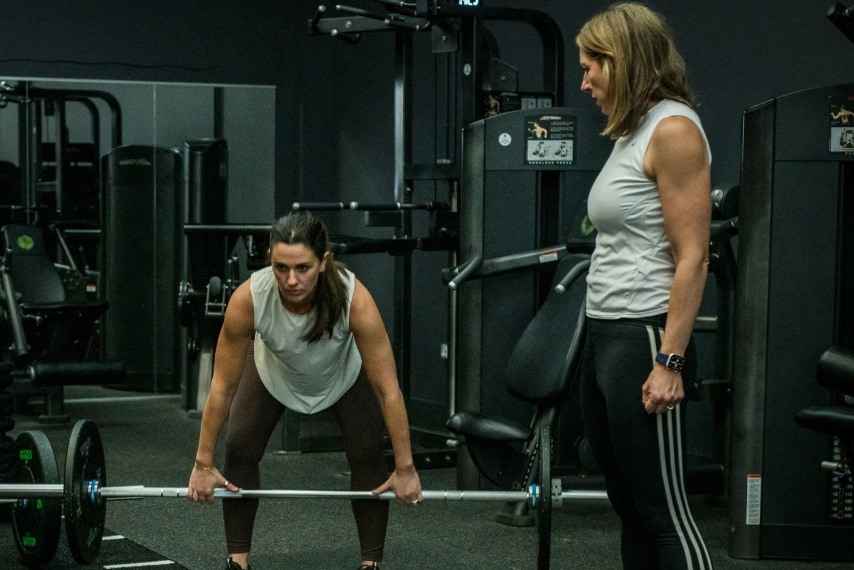Two women in a gym, one in a workout stance preparing to lift a barbell, the other standing nearby watching, with gym equipment in the background.