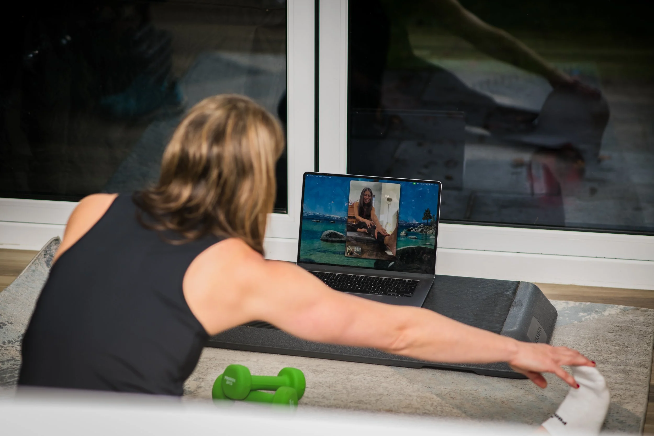 woman stretching on the floor during a video call, with a laptop in front of her showing a woman on the screen, a green dumbbell nearby, and a window behind her.