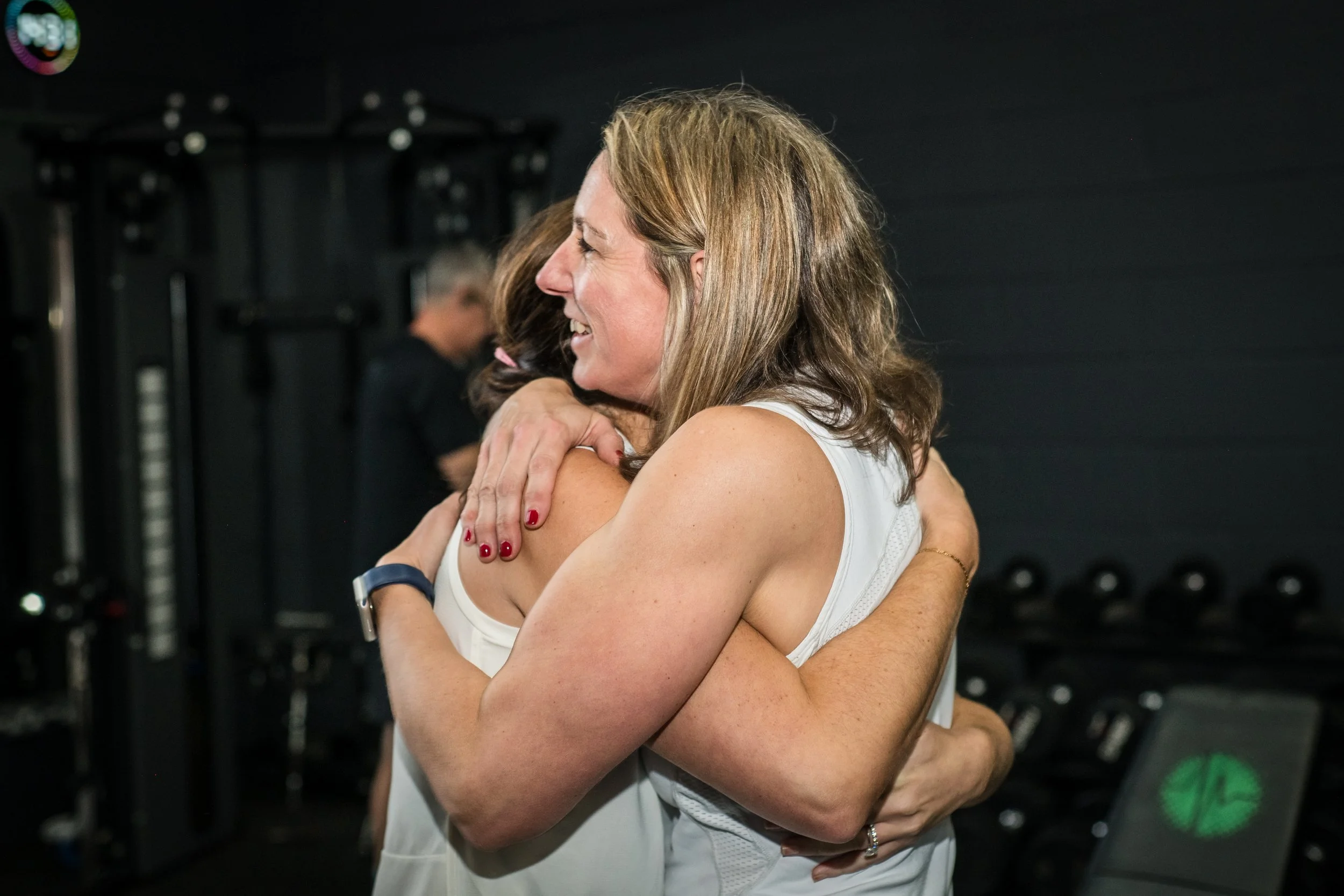 Two women hugging cheerfully in a gym, with workout equipment in the background.
