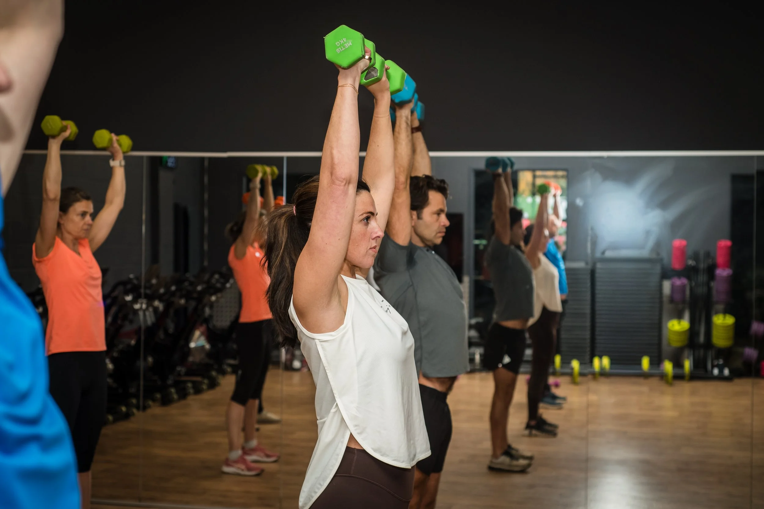 Group of people doing strength training with dumbbells in a gym, standing in front of a mirror.