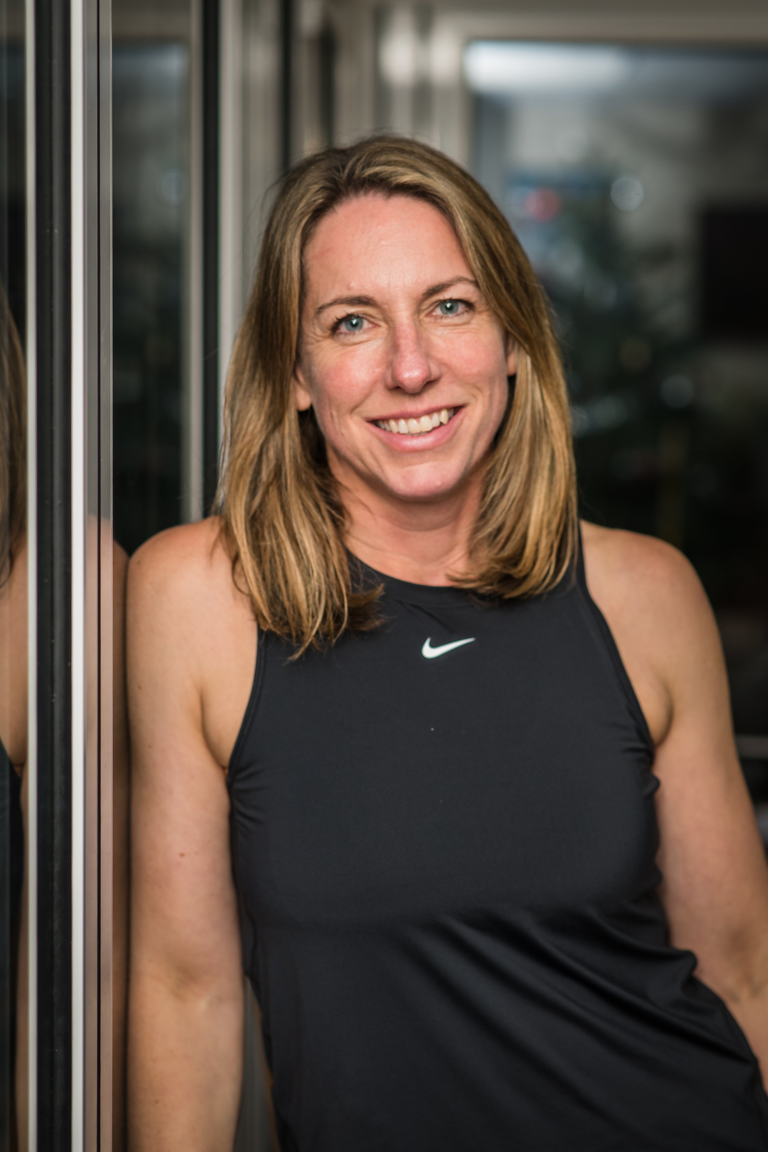 Smiling woman in black Nike athletic tank top leaning against a mirrored surface.