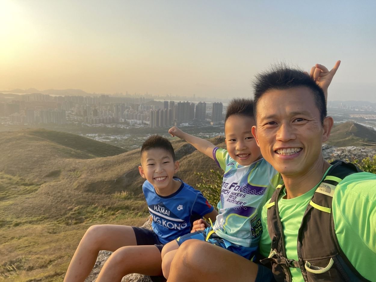 日落時分,晴朗的天空下,父親和兩個兒子在山頂上微笑著自拍,背景是城市的天際線。