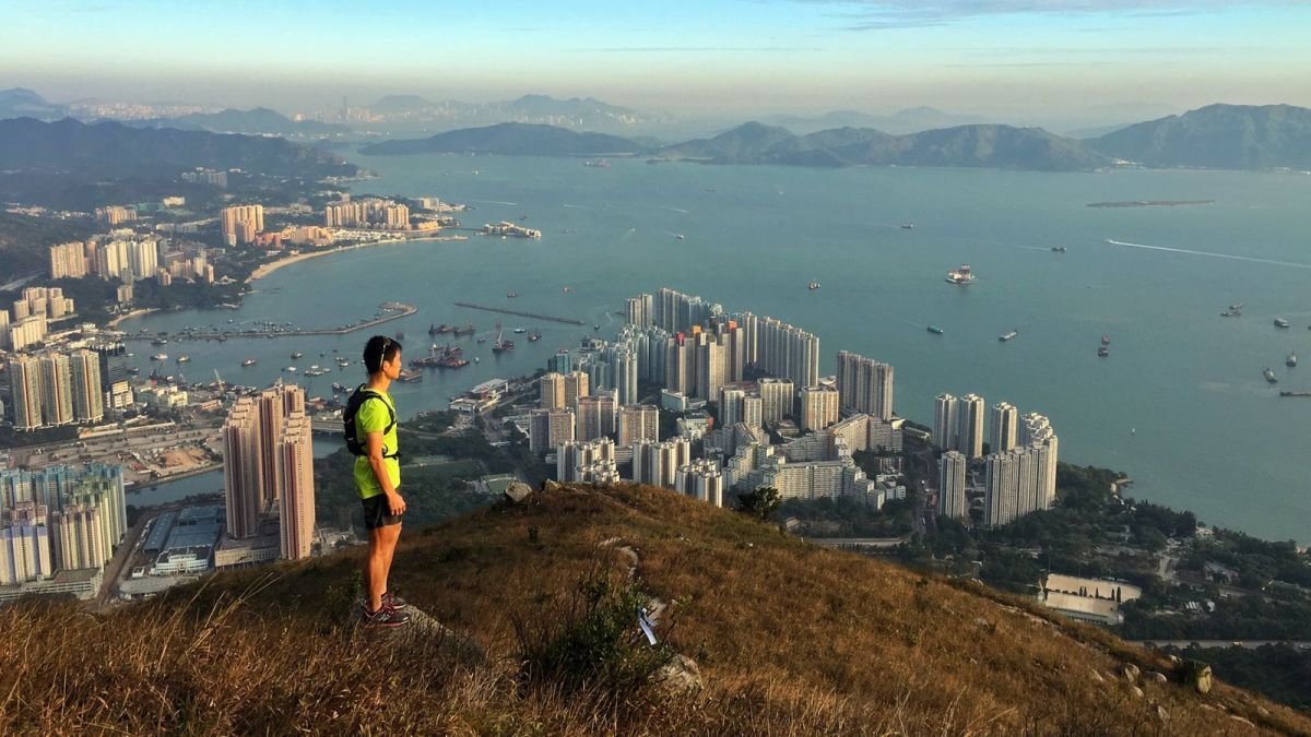 身穿運動服的人站在山上,俯瞰摩天大樓、港口和船隻等城市景觀,背景為群山。