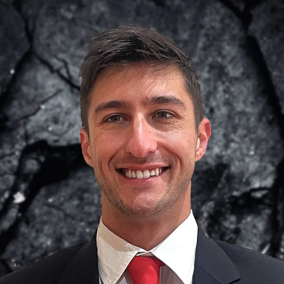 A young man in a suit and red tie smiling in front of a dark, textured background.