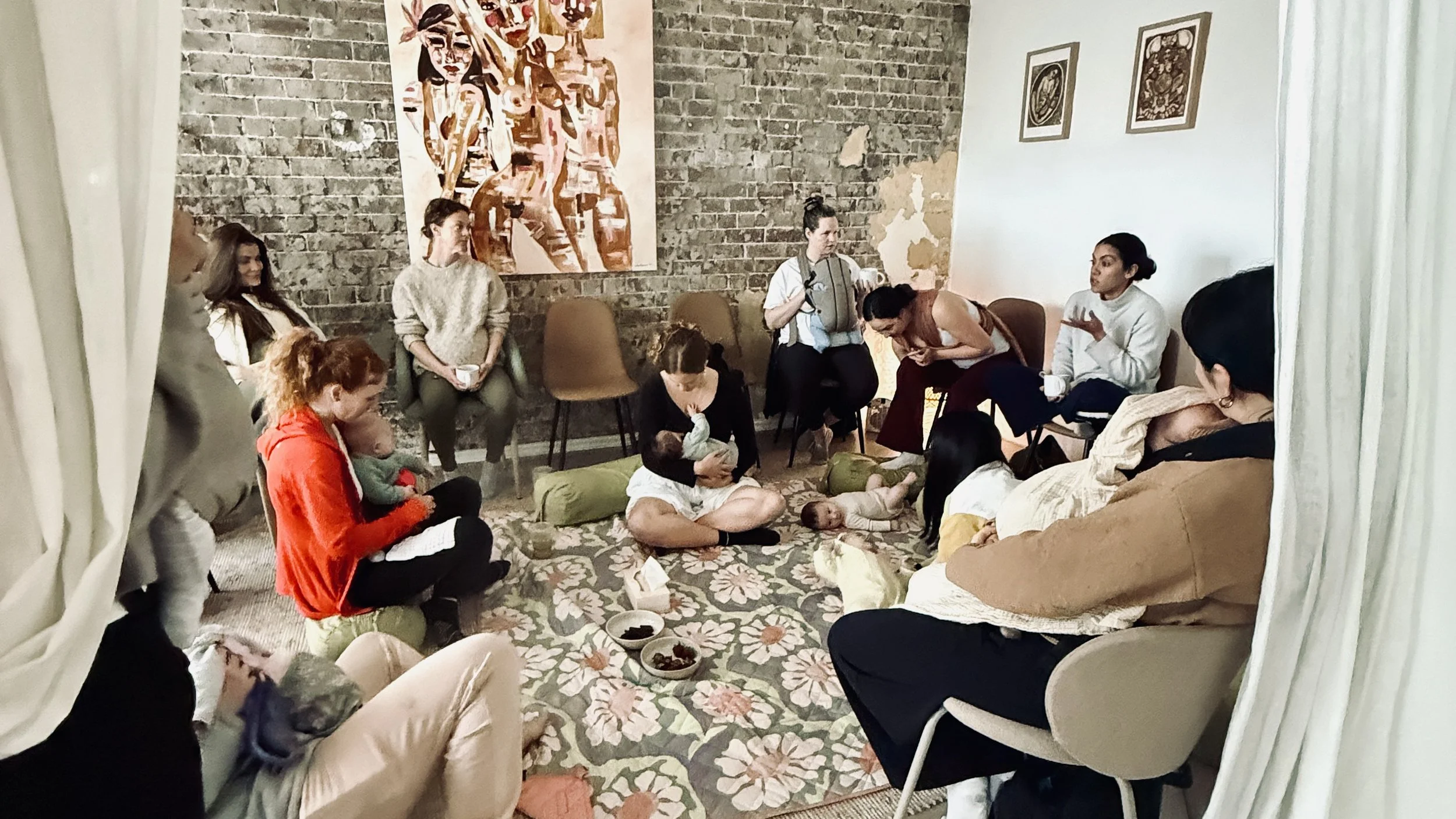 Group of women and babies gathered in a cozy room, some sitting on chairs and on the floor, participating in a breastfeeding support or parenting group. There are bowls of snacks or food on a floral rug, and the room has exposed brick walls with artwork hanging on them.