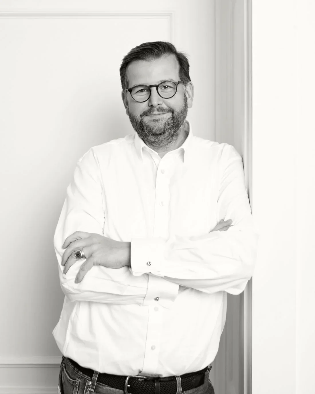 Schwarz-Weiß-Foto eines Mannes mit Brille, Bart und gepflegtem Haar, der lächelnd mit verschränkten Armen vor einer weißen Wand steht.