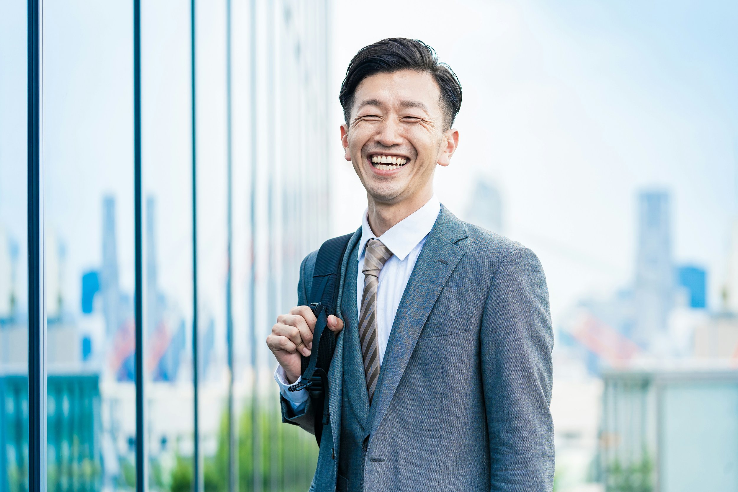 Smiling man in a gray suit with a backpack, standing outdoors near a reflective building, cityscape background.