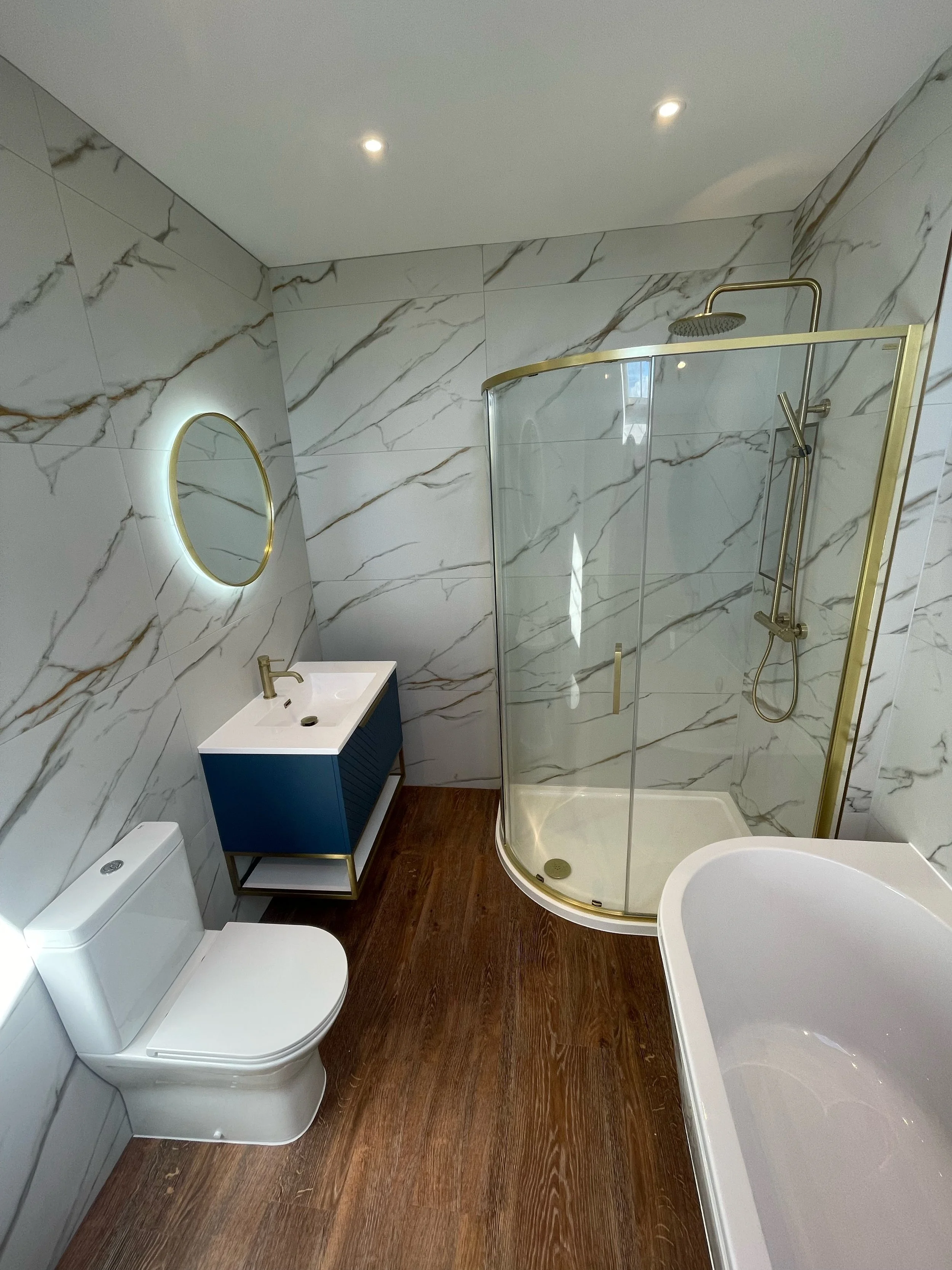 Modern bathroom with marble walls, circular mirror, blue vanity with gold faucet, glass shower with gold trim, toilet, and freestanding bathtub on wood floor.