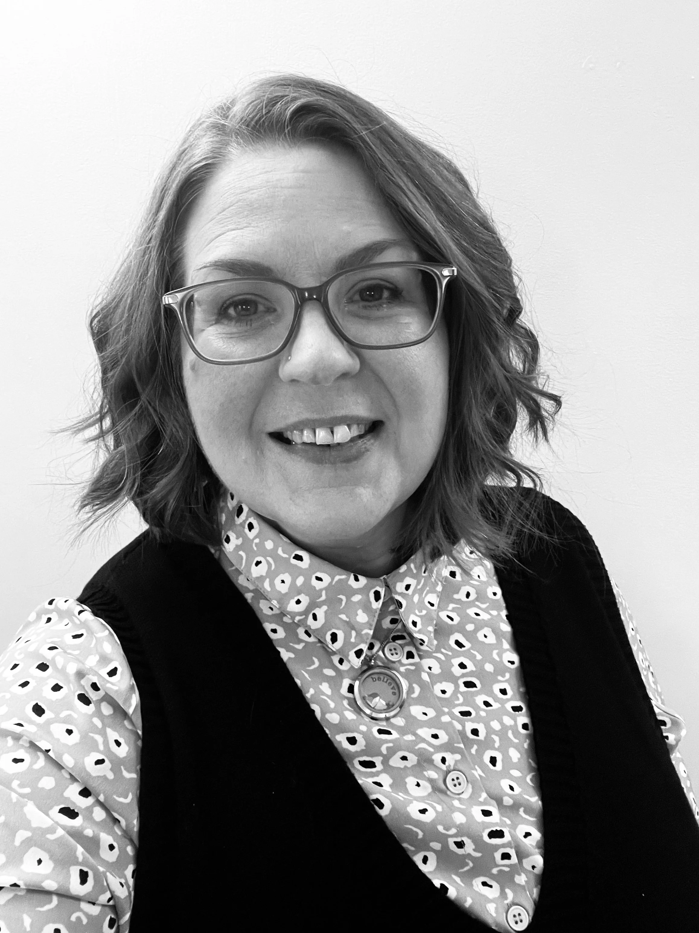 A woman with glasses and wavy hair smiling, wearing a patterned shirt and a dark vest, against a plain white wall.