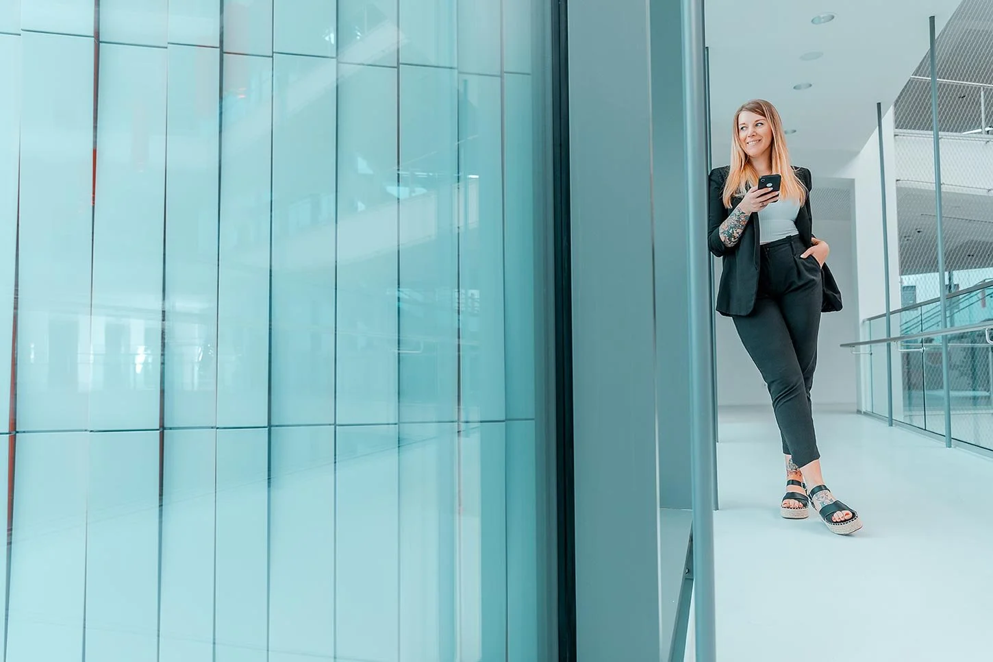 Businessfoto von Lisa Krieg Fotografie. Junge Frau in Businesskleidung steht in einem modernen Gebäude, lehnt an eine Wand aus Glas, hält ein Smartphone, lächelt und schaut nach oben.