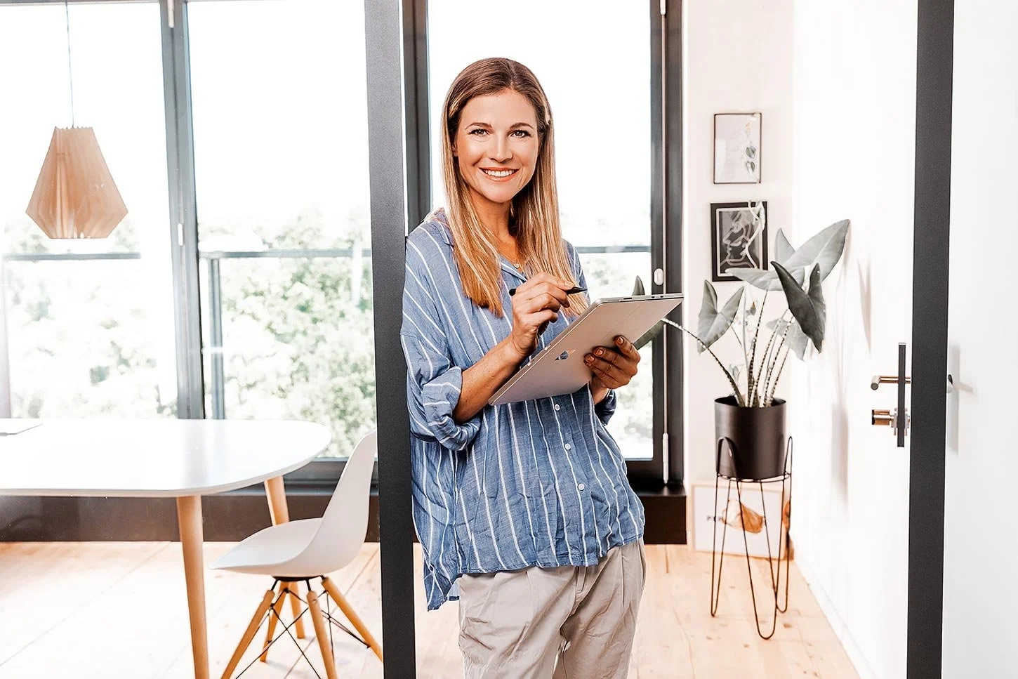 Businessfoto von Lisa Krieg Fotografie aus Mainz. Eine lächelnde Frau mit Notebook in einem modernen, hellen Büro, hält ein Notebook und einen Stift und lehnt sich an den Türrahmen.