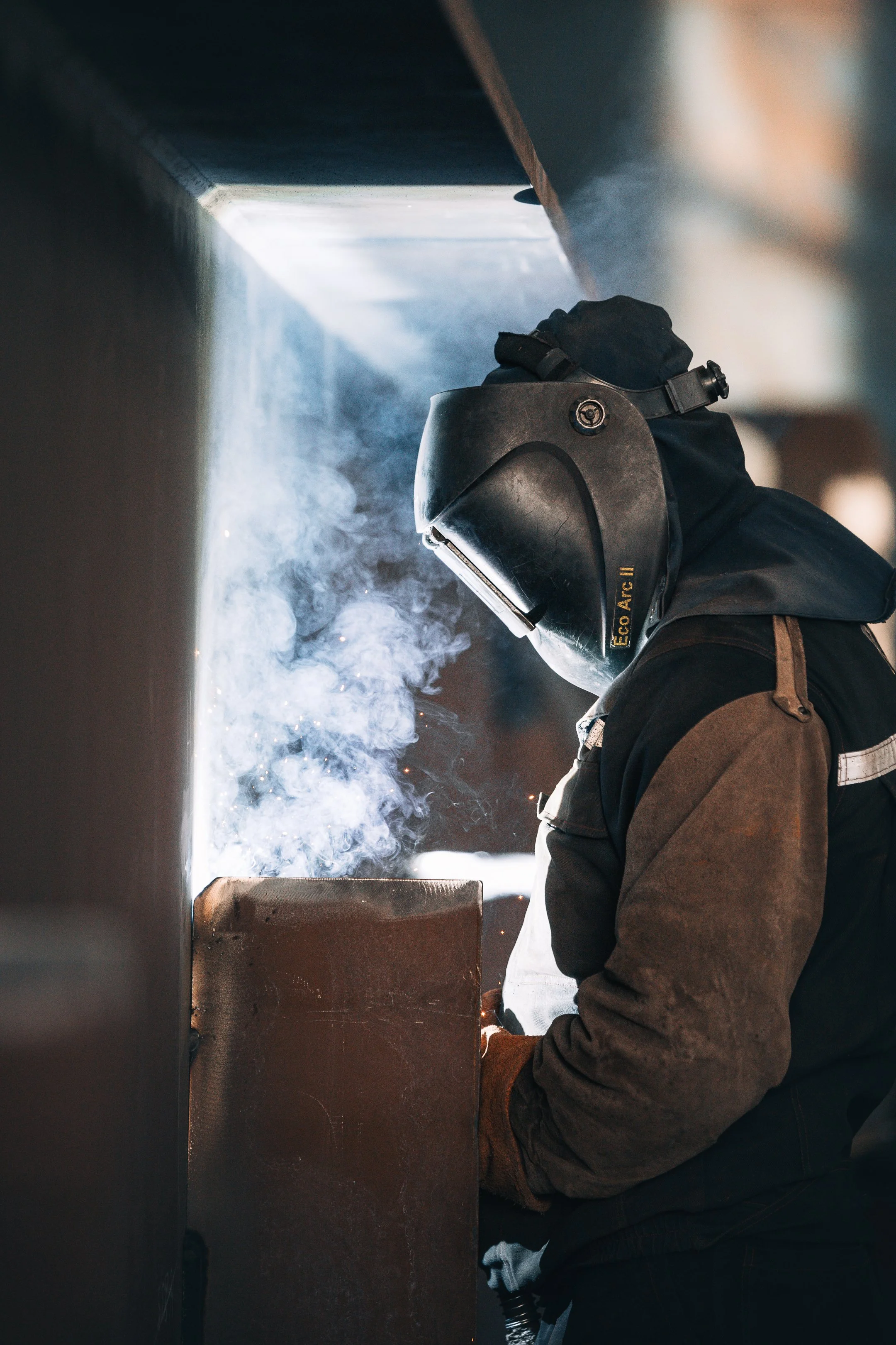 A person wearing a welding helmet and protective clothing is welding metal, with sparks and smoke visible.