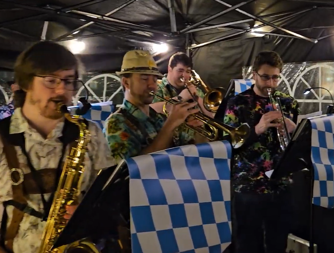 Four musicians playing brass instruments at a nighttime outdoor event, decorated with blue and white checkered flags.