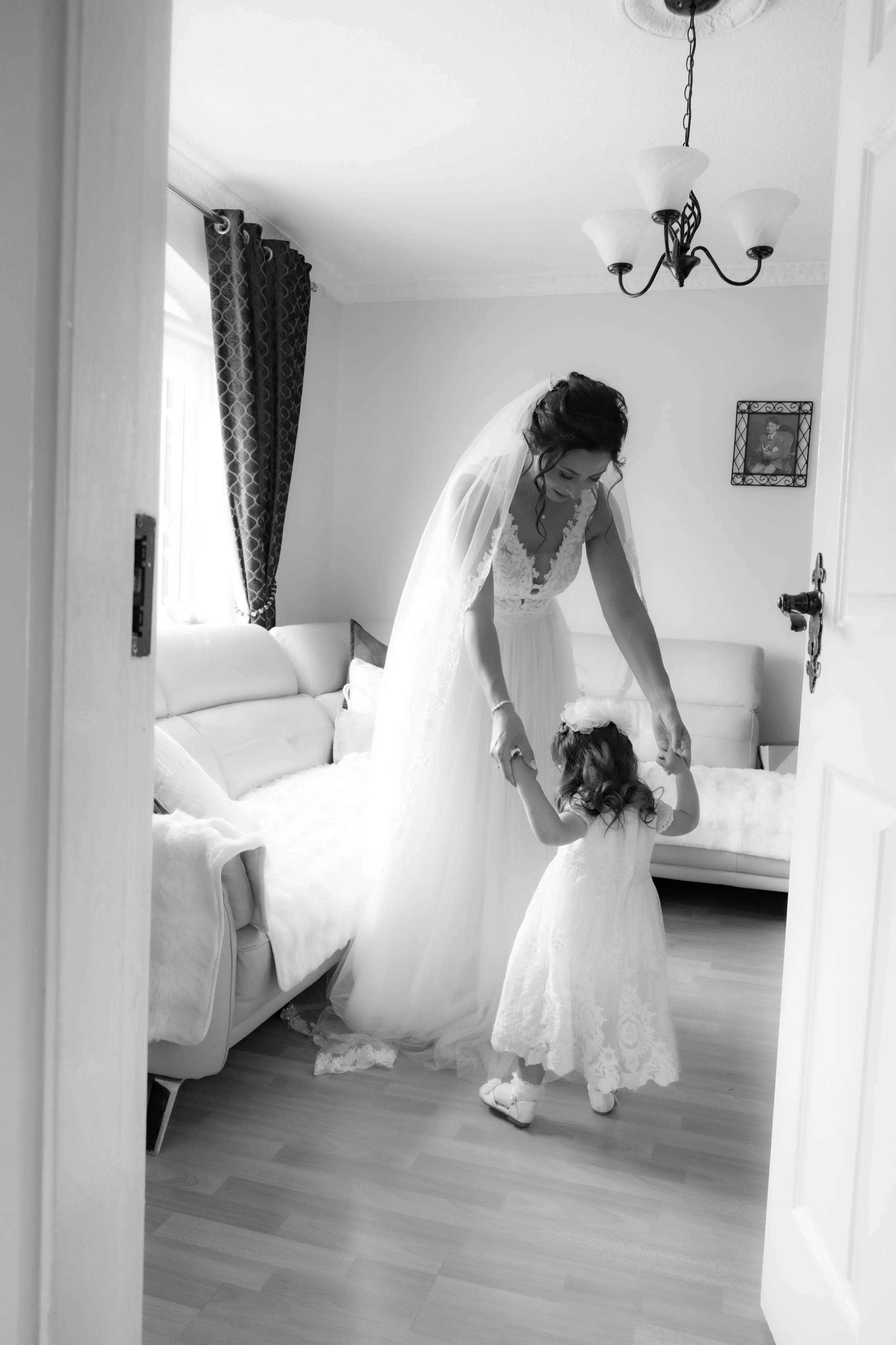 A bride and a young girl are holding hands inside a living room, possibly practicing a dance or having a moment before a wedding. The bride is wearing a wedding dress and veil, and the girl is in a matching dress.
