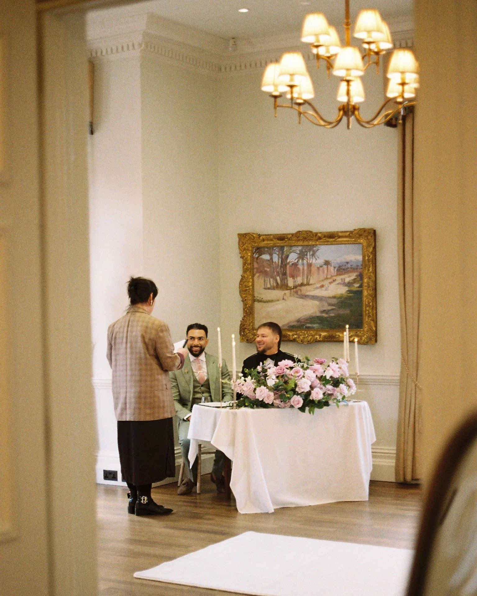 A wedding ceremony taking place indoors with a table decorated with pink flowers in the foreground. Two men are seated at the table, smiling, in front of a large framed landscape painting. A woman in a plaid blazer is standing and reading from a pape