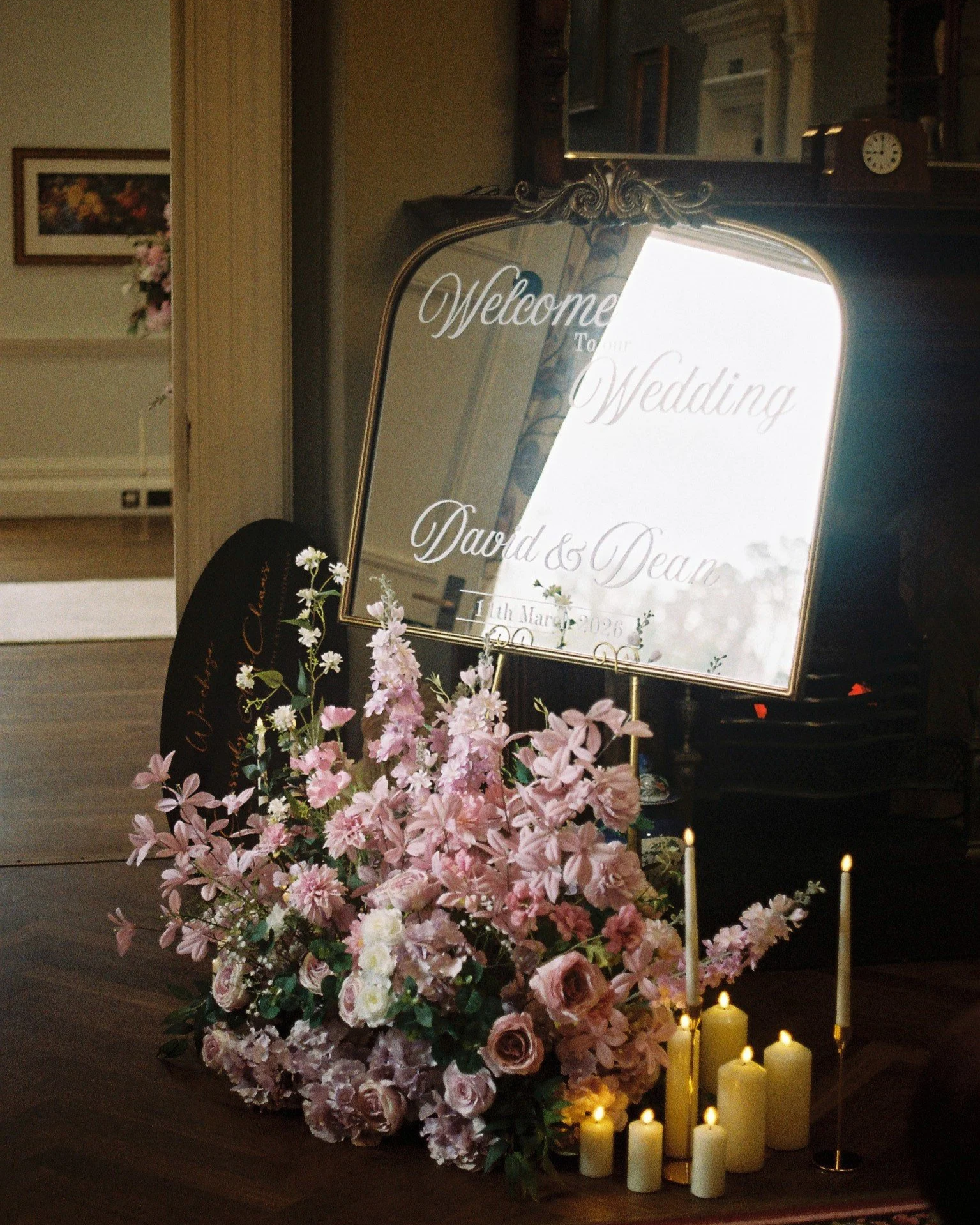 A decorative wedding welcome sign with white text on a mirror, surrounded by a large arrangement of pink and white flowers, with candles at the base.