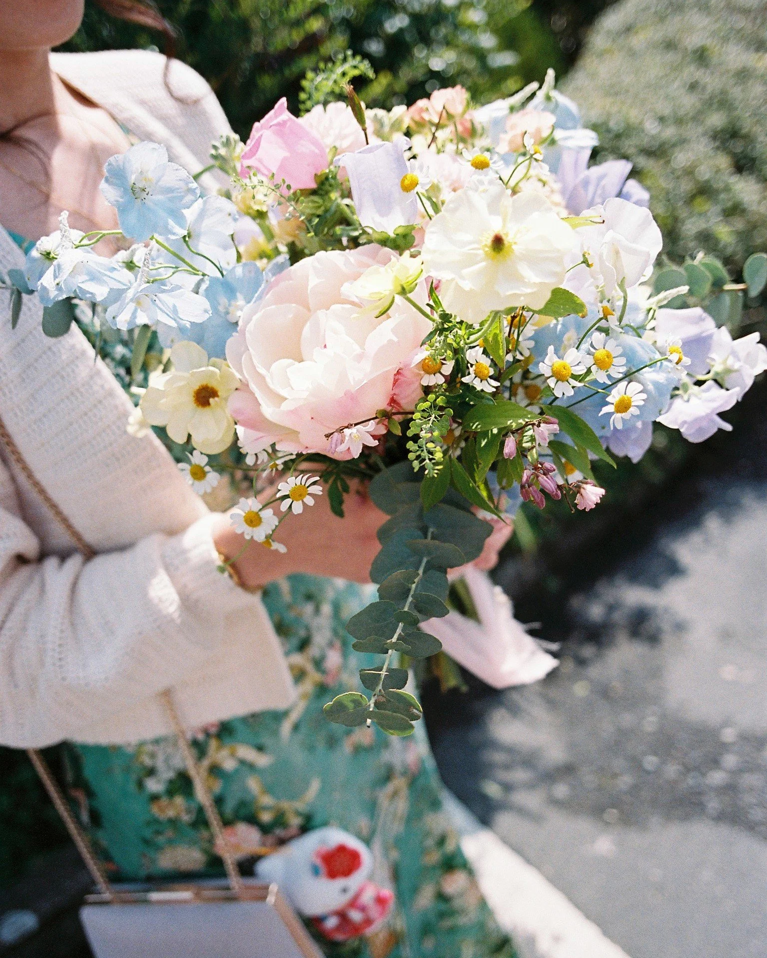 A person holding a colorful bouquet of flowers including pink, white, light blue, and small yellow flowers with green leaves. The person's face is not visible and they are wearing a cream-colored sweater.