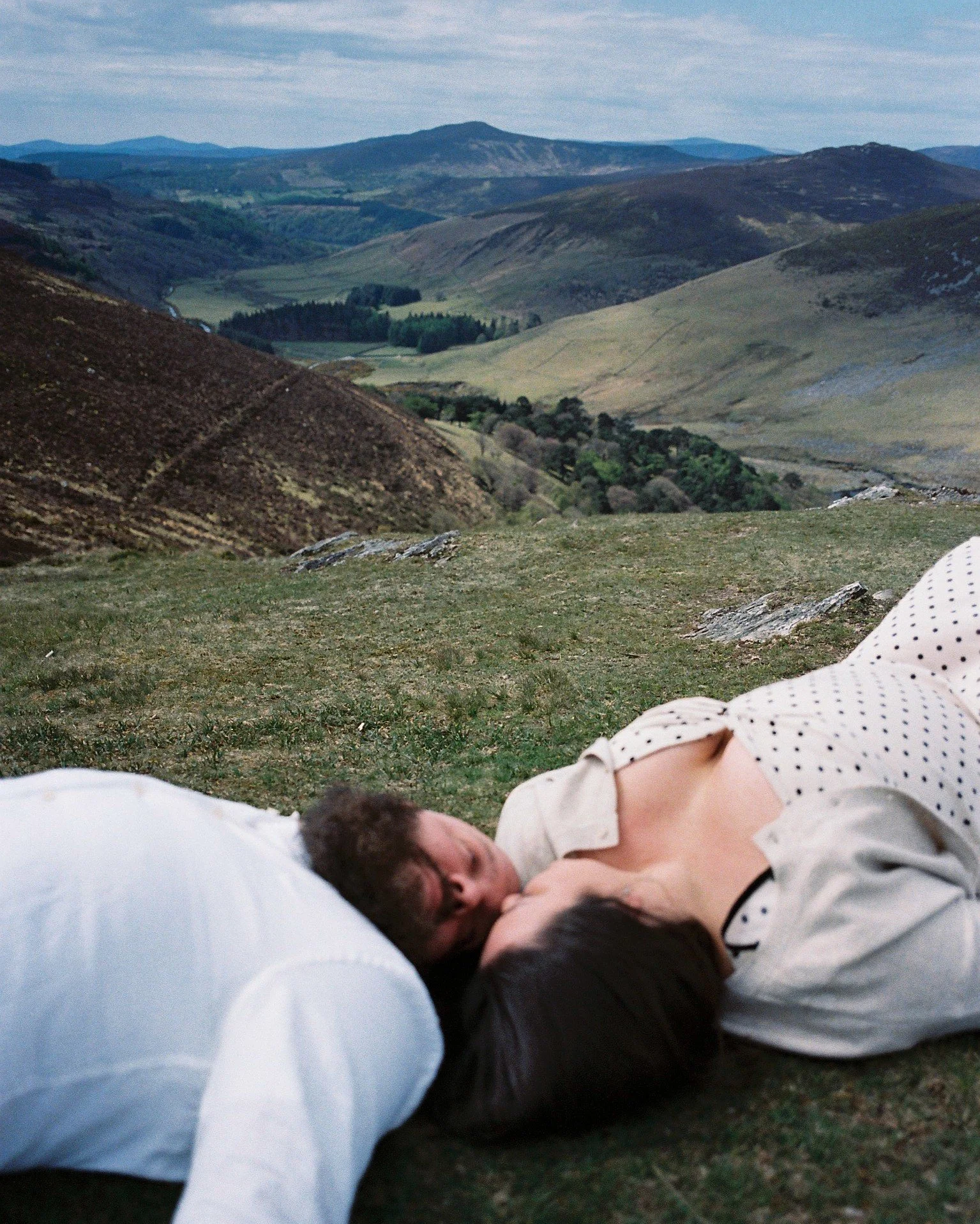 A person lying on the grass with mountains and rolling hills in the background.