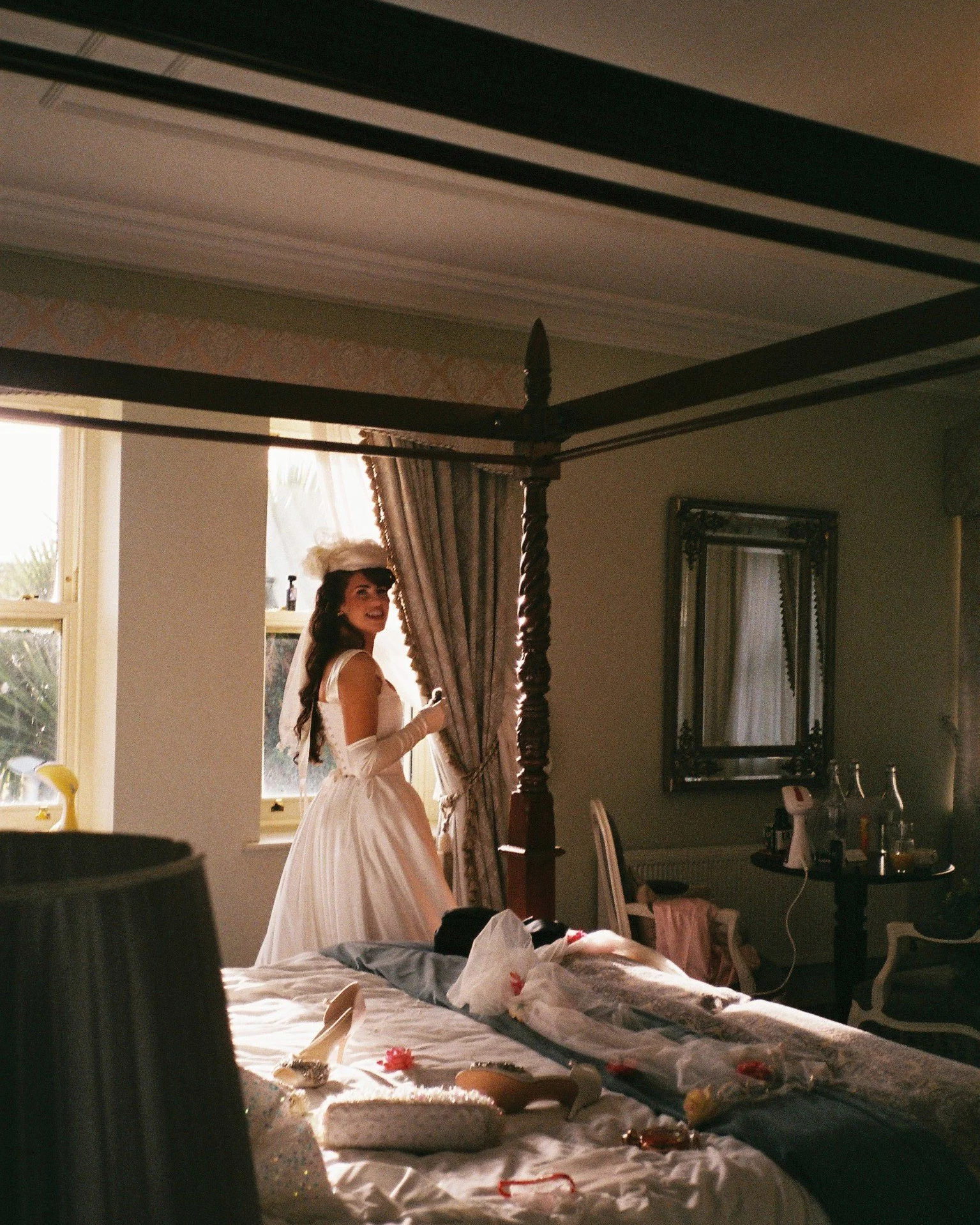 A woman dressed in a vintage white dress with long gloves, standing in a sunlit room near a window. She is smiling and looking to her left, with long dark hair and a feathered headpiece. The room has dark wood furniture, including a four-poster bed, 