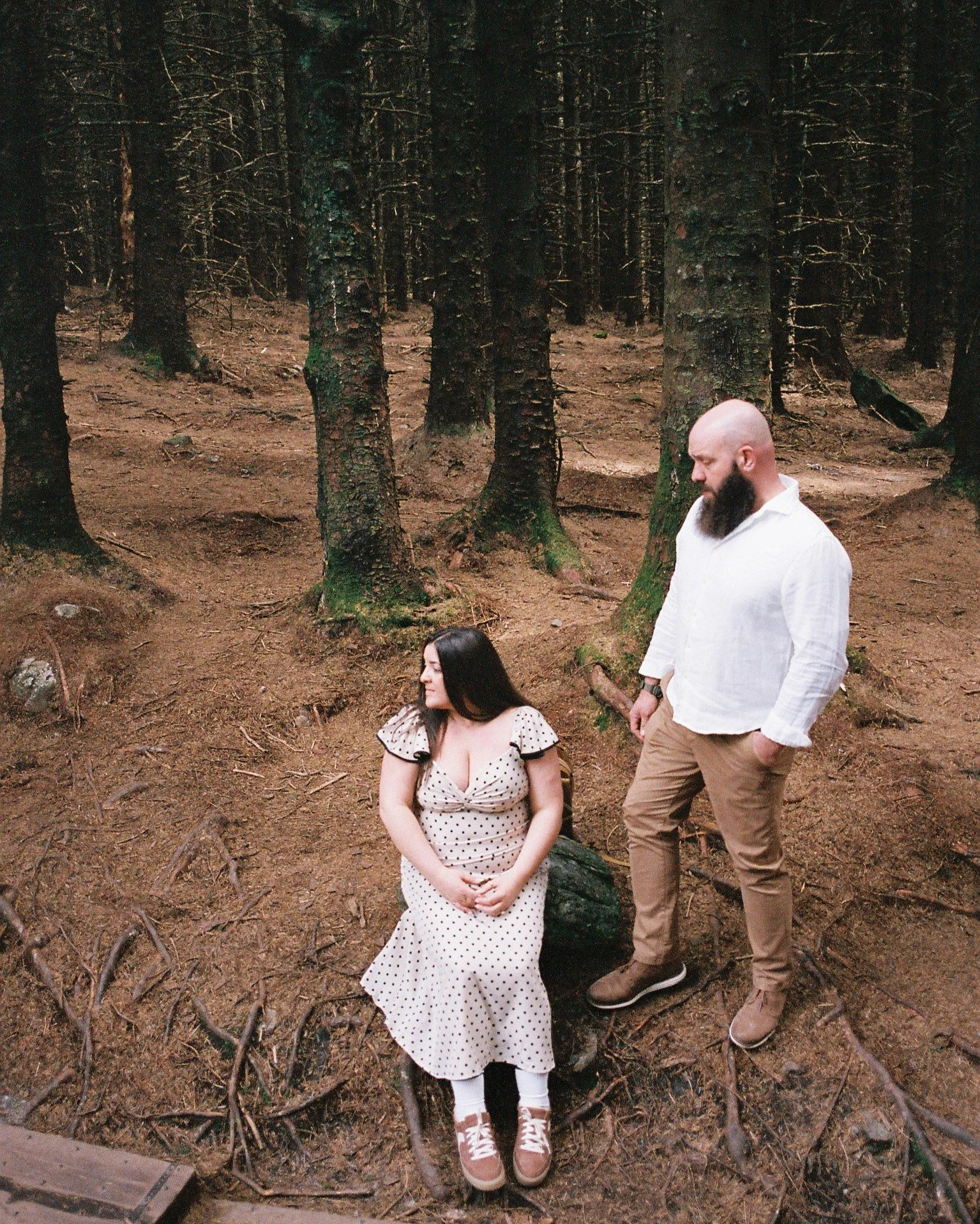 A woman sitting on a tree stump in a forest, wearing a white polka dot dress, and a man with a bald head and beard standing beside her, wearing a white shirt and brown pants.