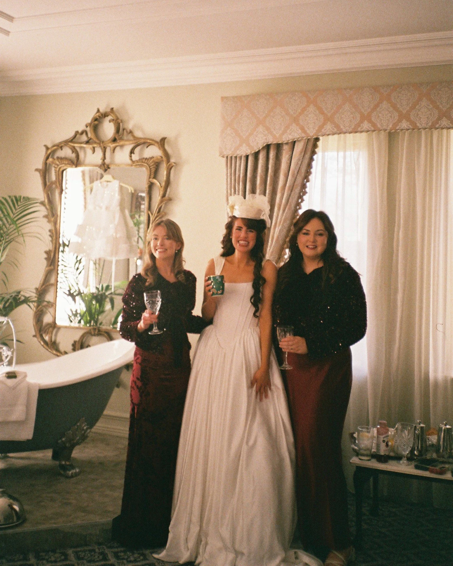Three women at a party or celebration, one wearing a wedding dress and a veil, holding a mug, and the other two in dark dresses holding wine glasses, standing in a room with vintage decor, a large mirror, and a clawfoot bathtub.