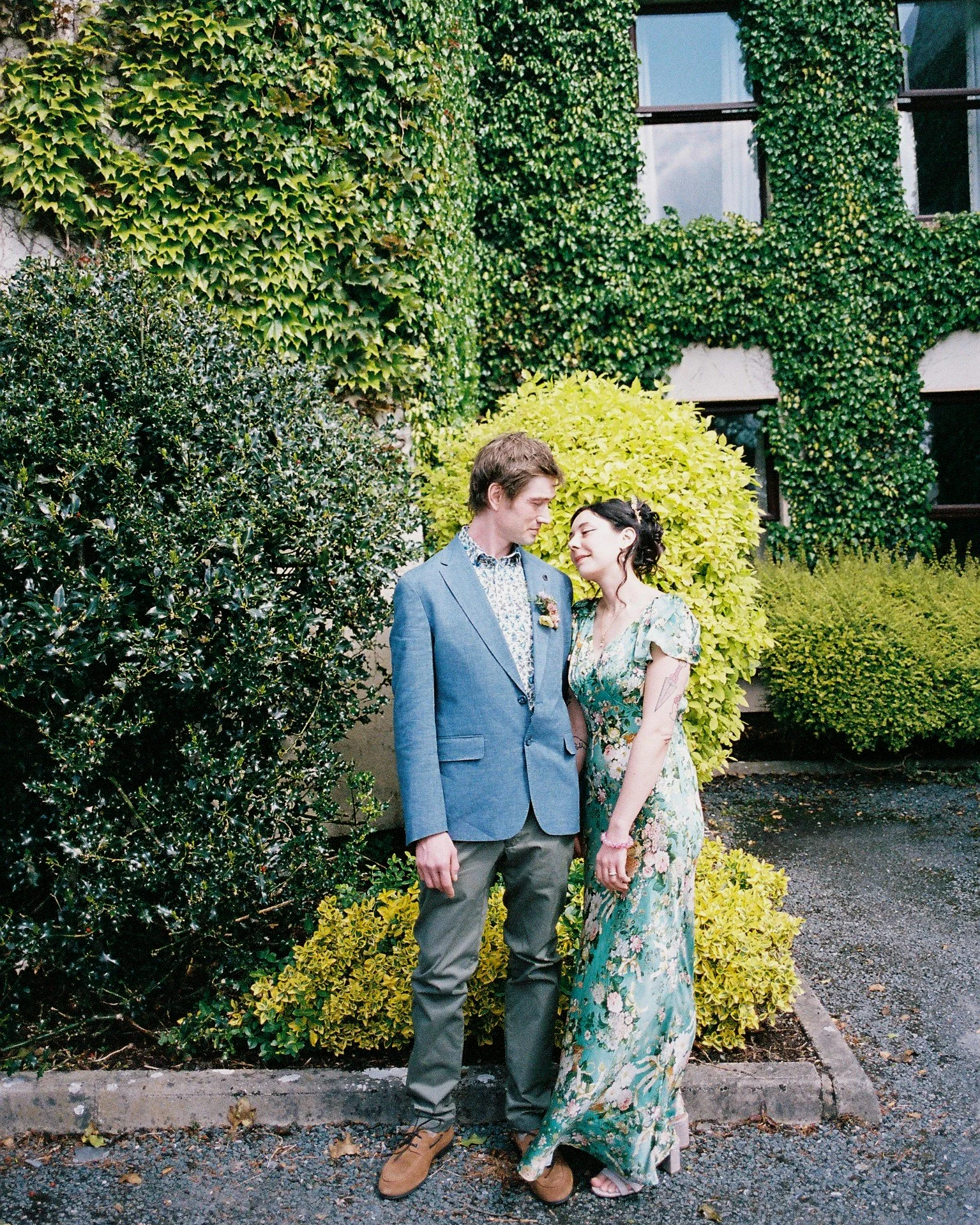 A young couple standing close together outdoors in front of a green, leafy wall with windows. The man has brown hair, wearing a light blue suit jacket, a patterned shirt, gray pants, and brown shoes. The woman has black hair, wearing a floral dress w