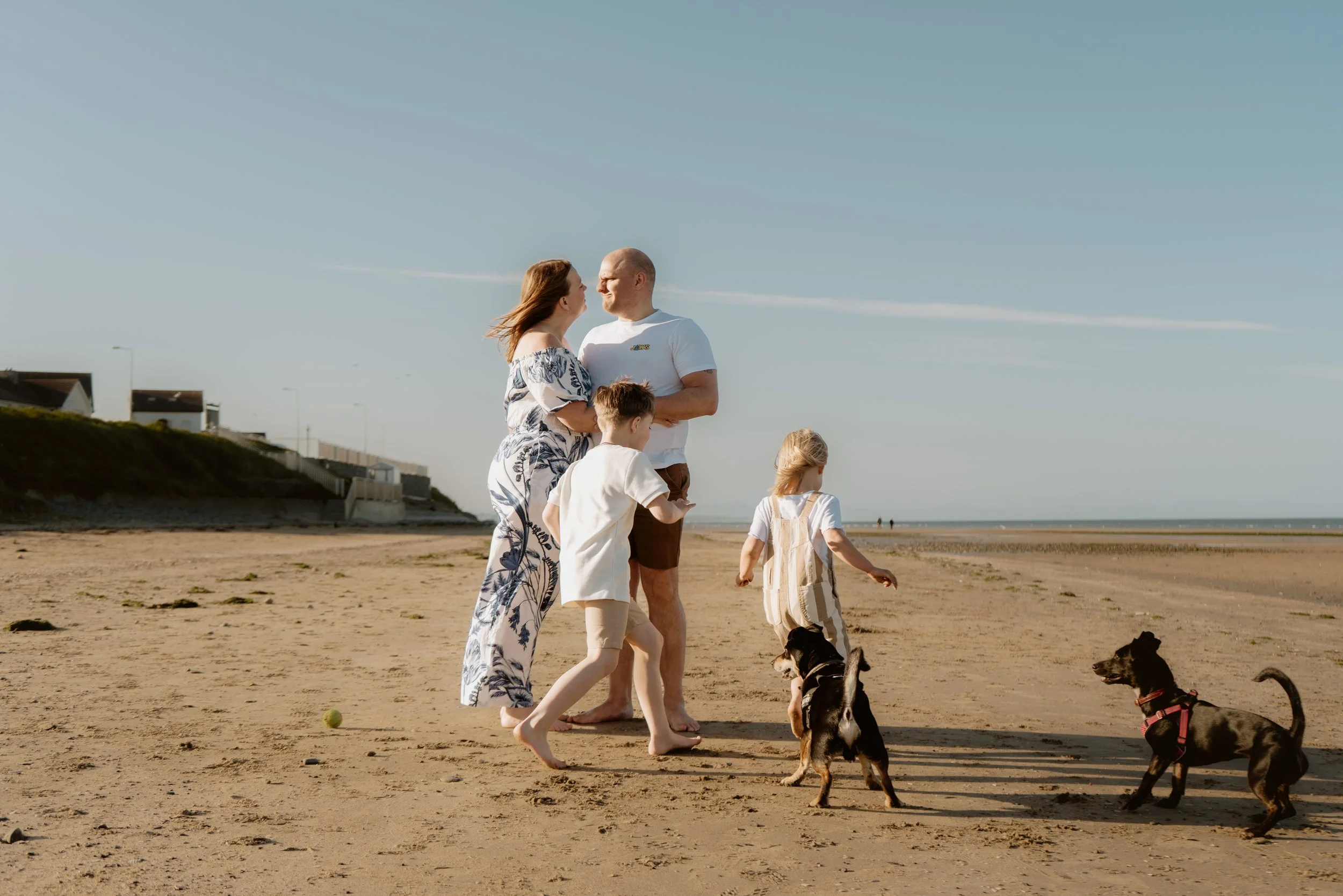 Family consisting of adults, two children, and dogs enjoying a walk on the beach under a clear sky.