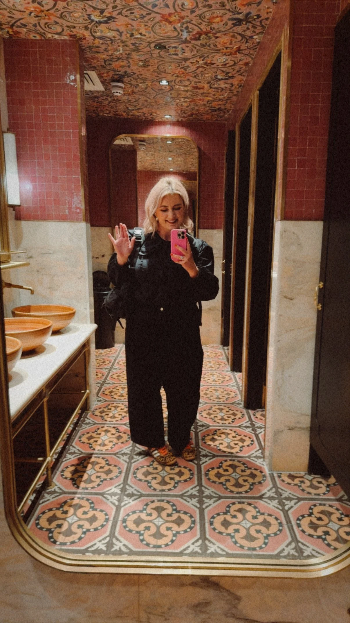 A woman with blonde hair taking a selfie in a decorative bathroom mirror. She is smiling and waving, wearing dark clothing and carrying a black backpack.