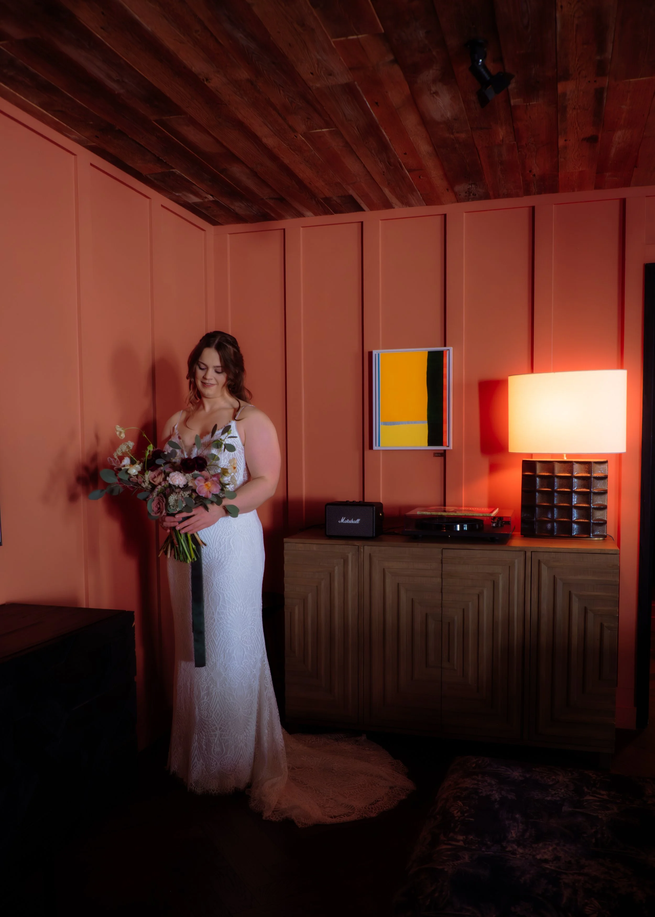 A woman in a white dress holding a bouquet of flowers standing in a warmly lit room with wood-paneled walls, a lamp, a colorful abstract painting, and a turntable on a wooden cabinet.