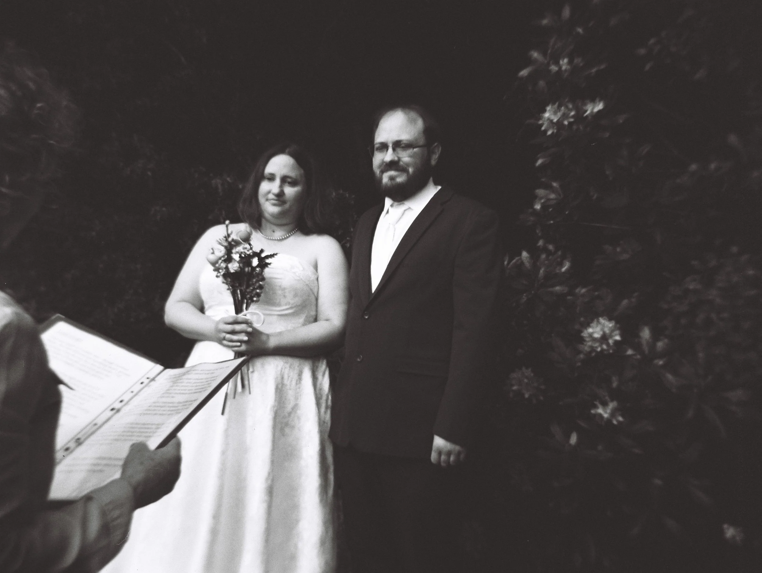 Black and white photo of a bride and groom at their wedding, standing outdoors with trees and foliage in the background. The bride is holding a bouquet of flowers, and the groom is wearing glasses and a suit.