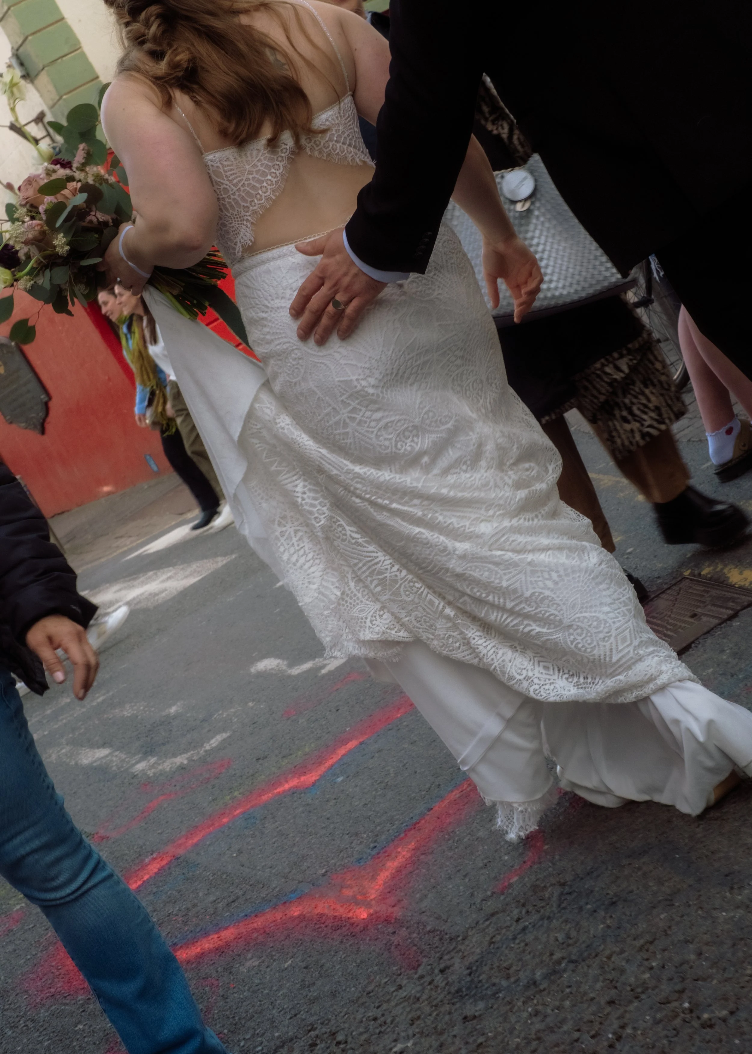Close-up of a bride in a white lace wedding dress holding a bouquet, surrounded by people on a street.
