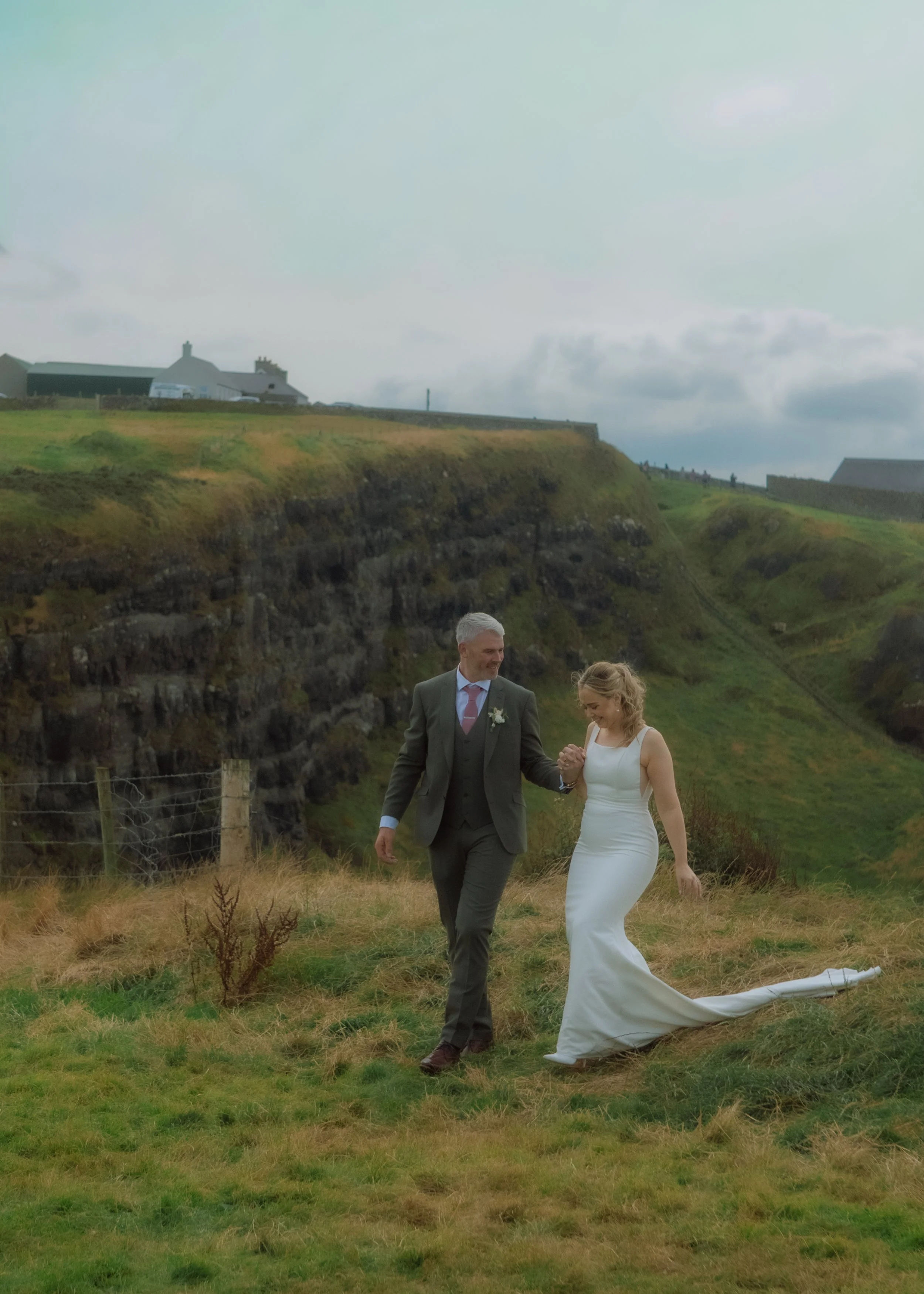 A bride and groom holding hands and walking on grass in a hilly landscape, with houses and a cloudy sky in the background.