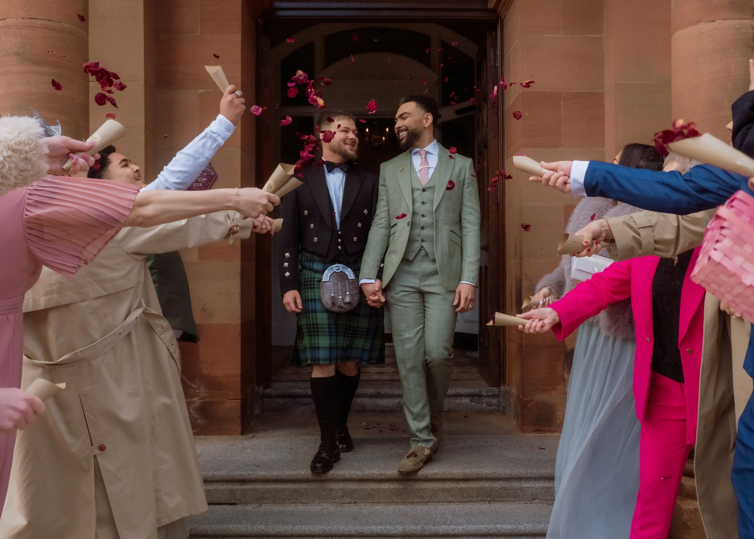 Couple walking out of a building holding hands while friends celebrate with confetti and paper tubes.